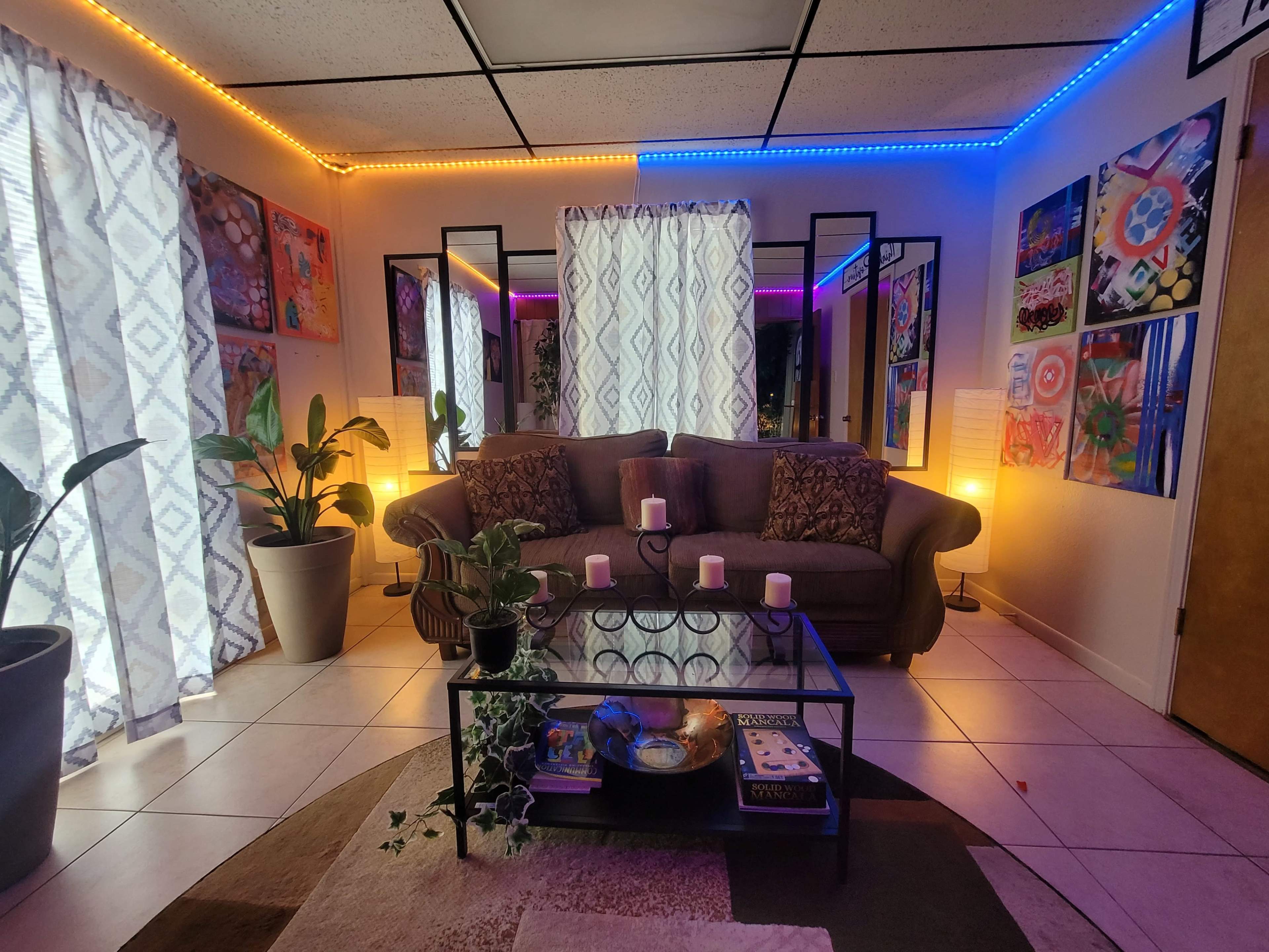 A living room with a gray sofa, a glass coffee table, and colorful LED lights illuminating the walls, surrounded by artwork and plants.