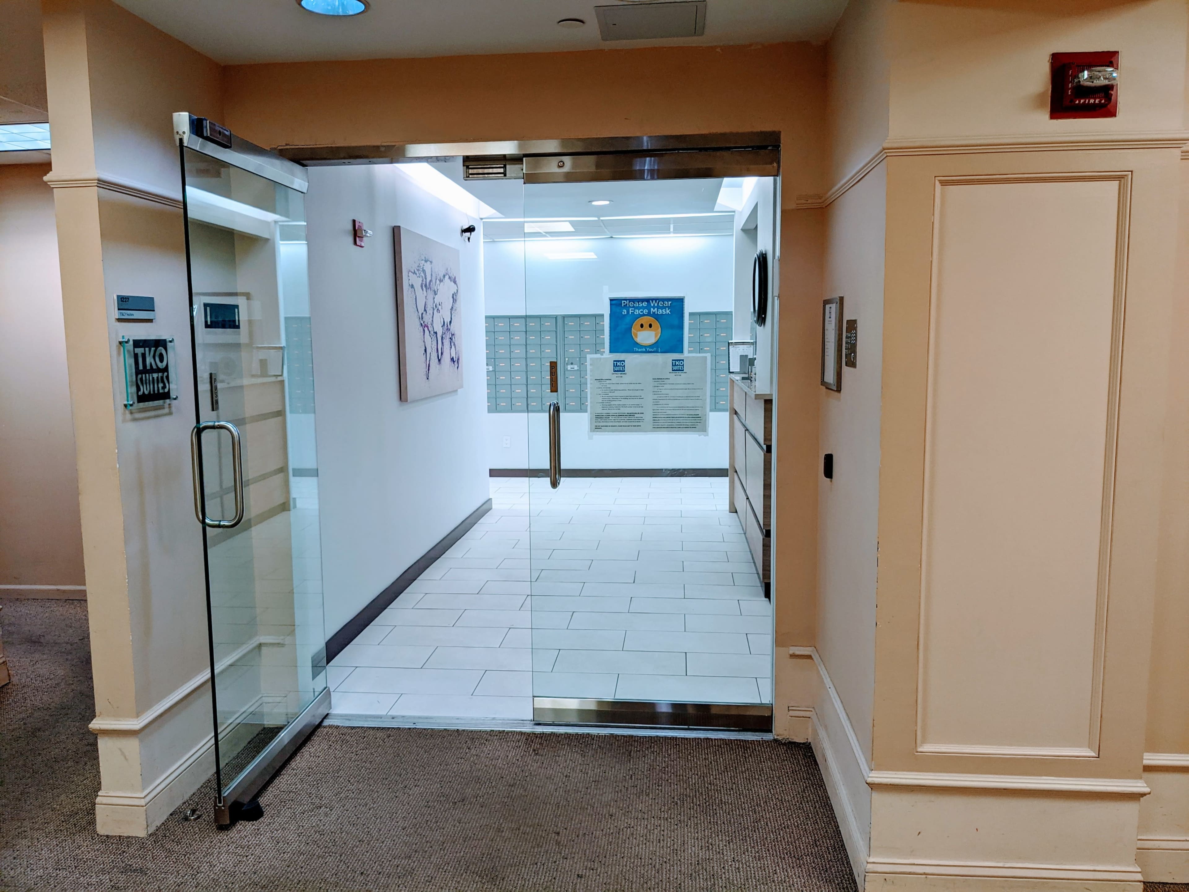 The image shows a glass entryway leading into a hallway with mailboxes and a bright interior space.