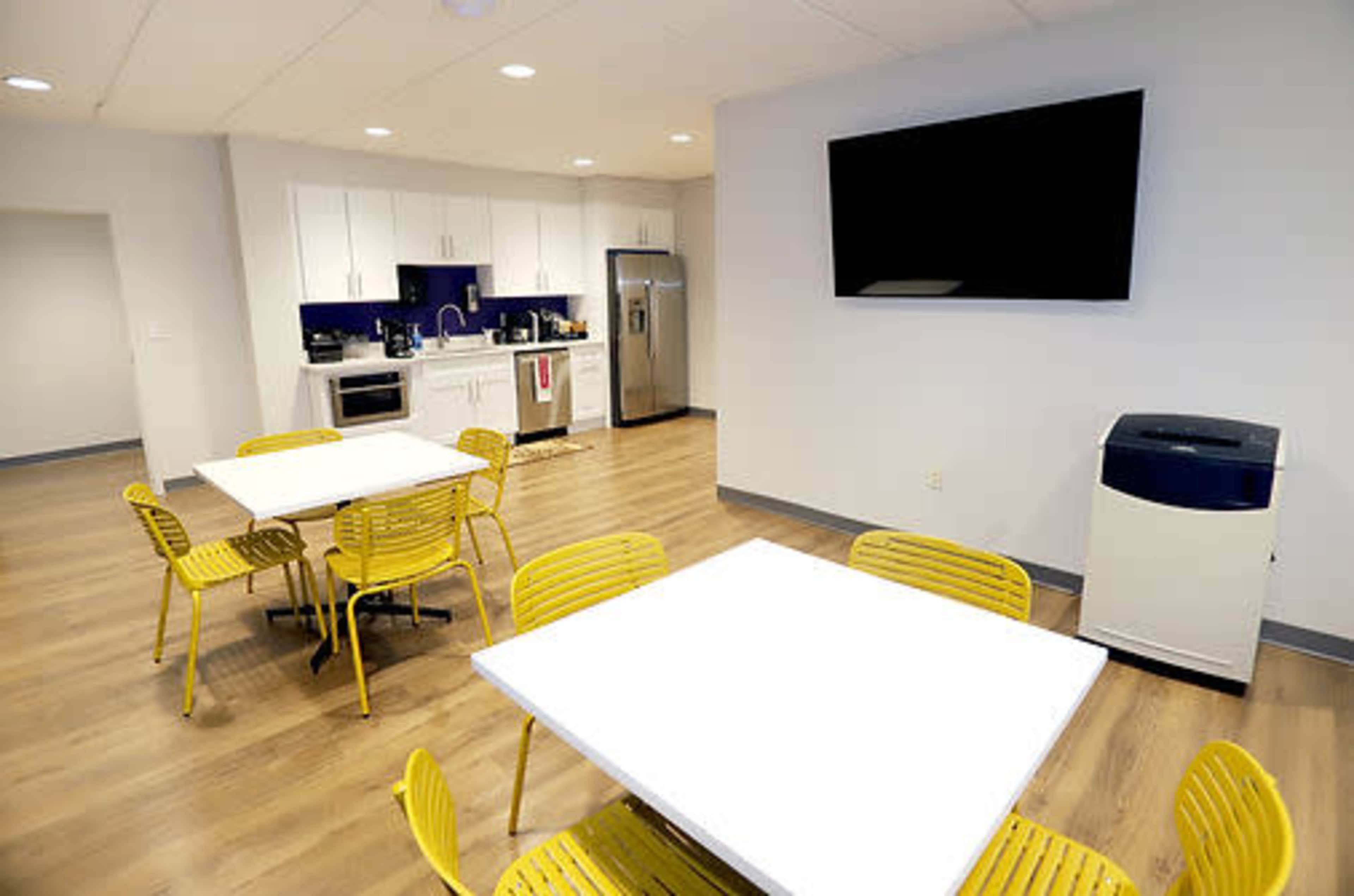 The image shows a modern kitchen and dining area with a refrigerator, stove, and a large television, complemented by yellow chairs and tables on a wood floor.
