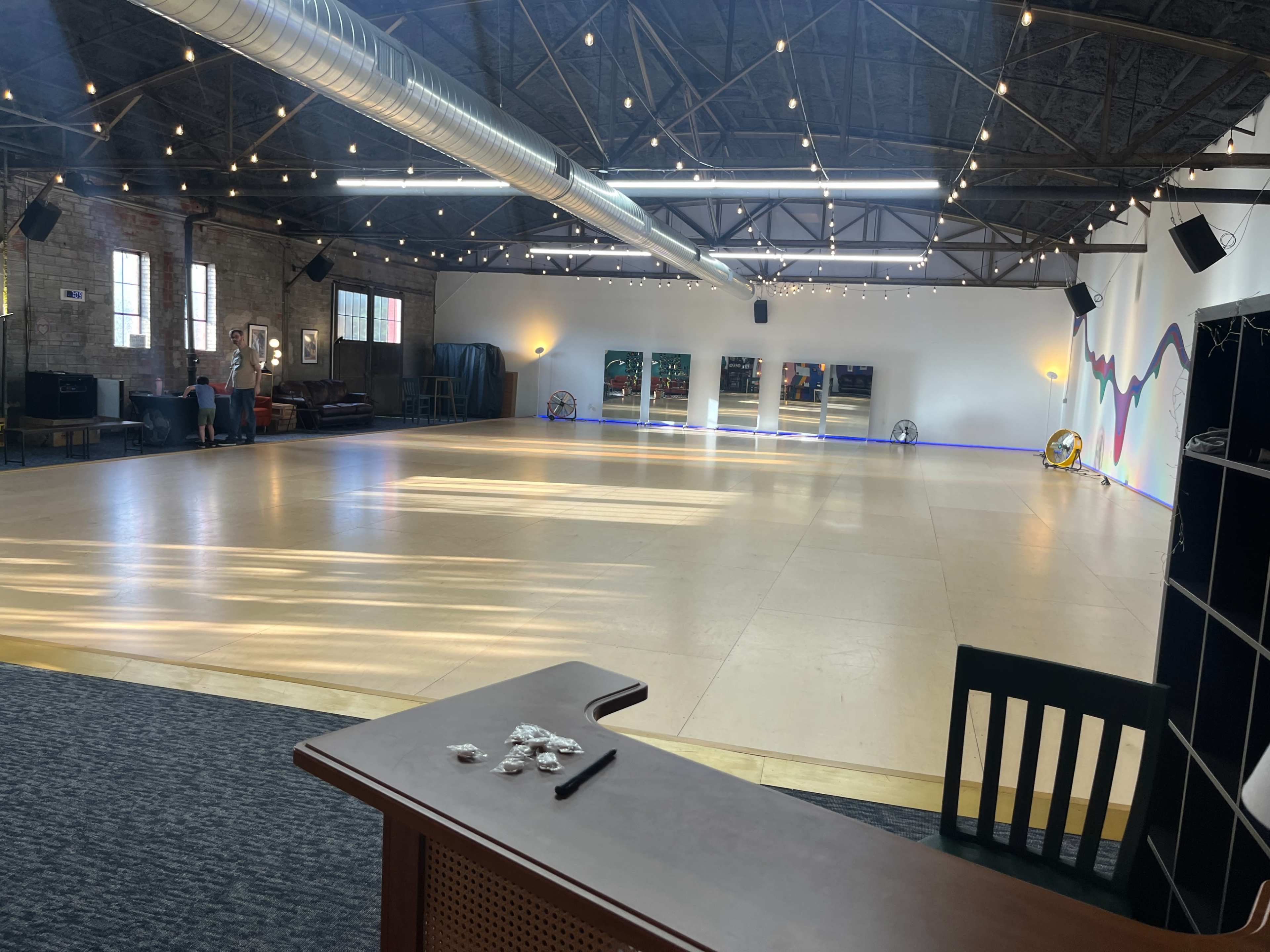 The image shows a spacious dance studio with wooden flooring, large mirrors on one wall, and a variety of seating areas along the sides, illuminated by overhead string lights.