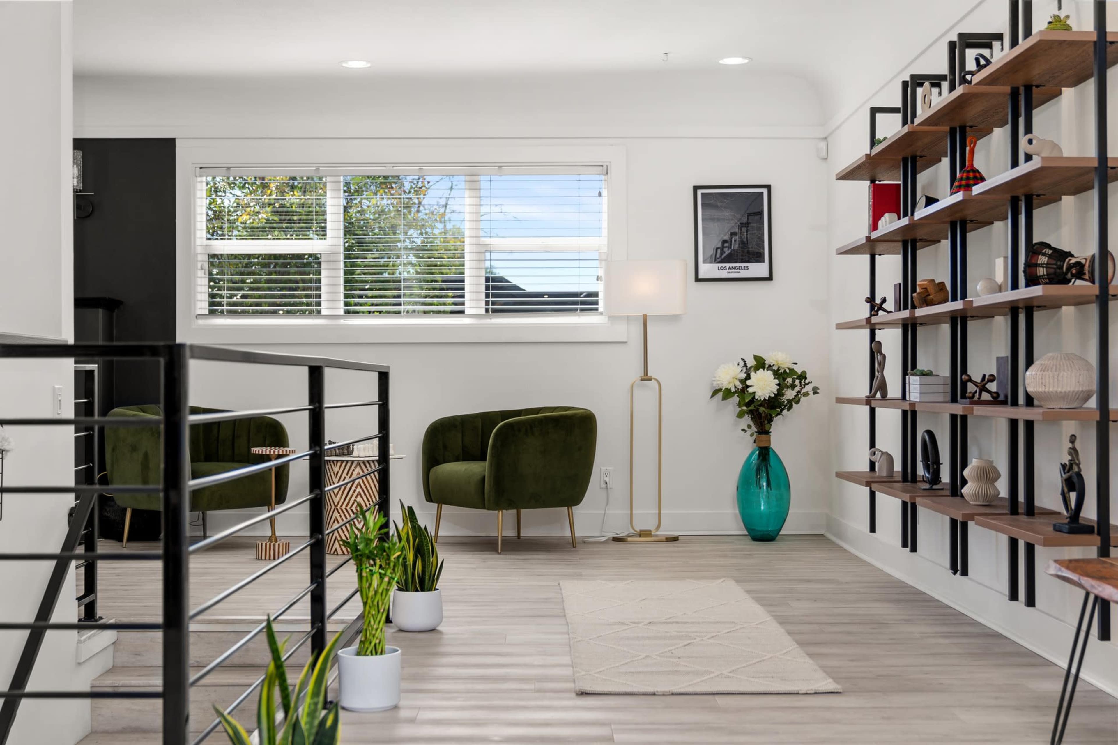 A modern interior features a green armchair, a floor lamp, a bookshelf with decorative items, and potted plants along a light-colored floor.