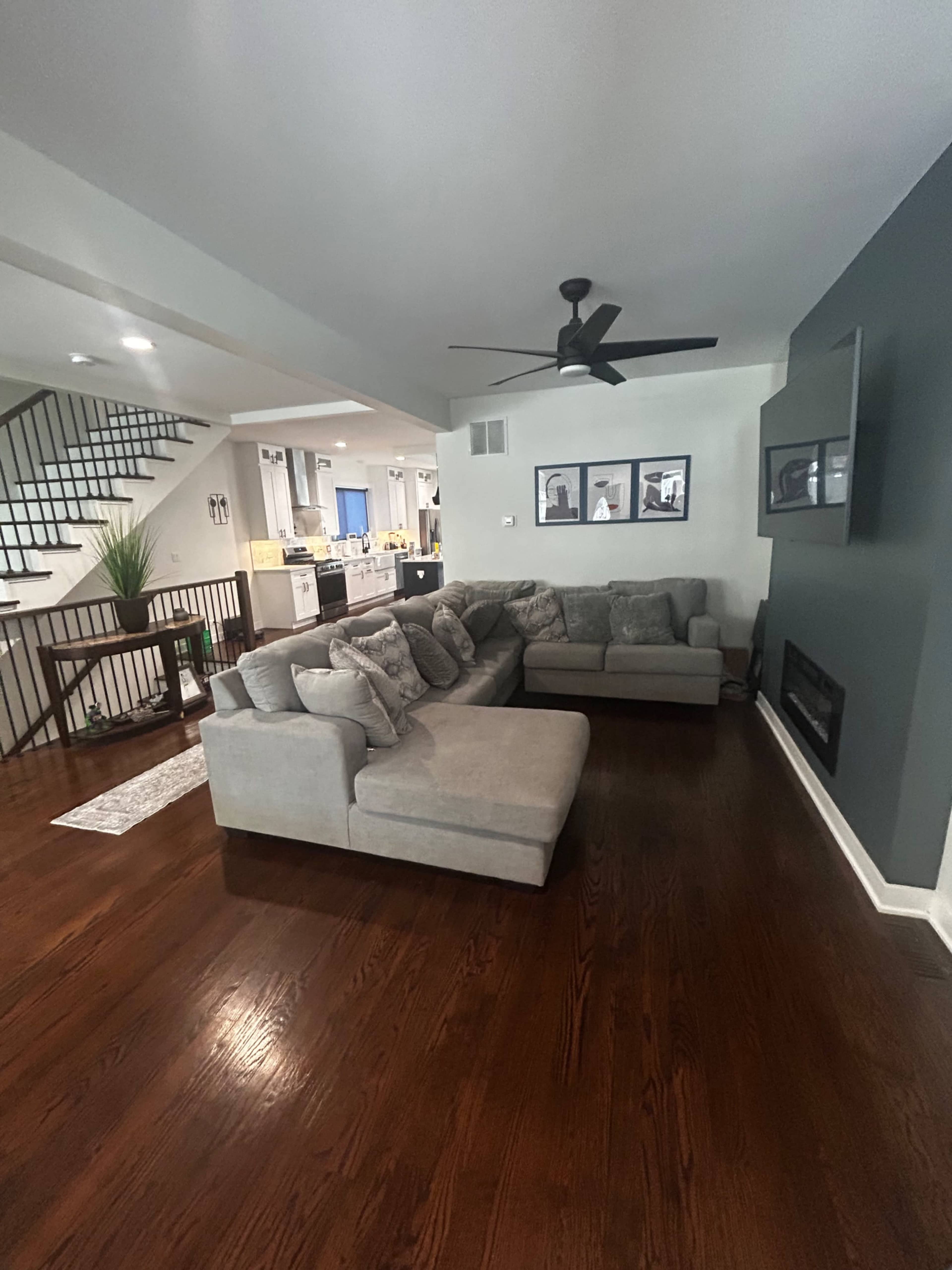 The image shows a modern living room featuring a large gray sectional sofa, a wall-mounted television, and a staircase in the background.