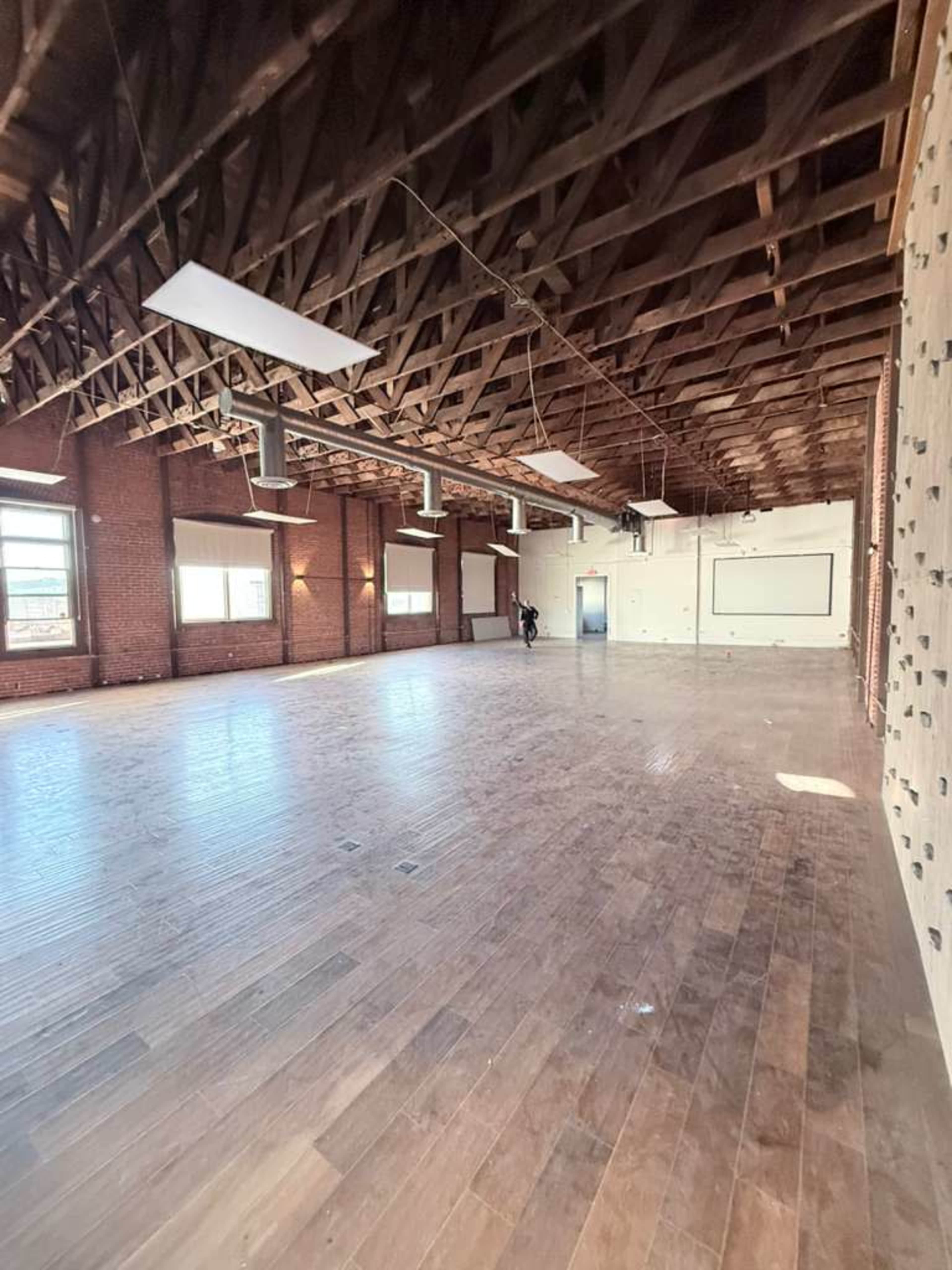 The image shows a spacious, empty room with wooden flooring, exposed beams, and large windows along one side.