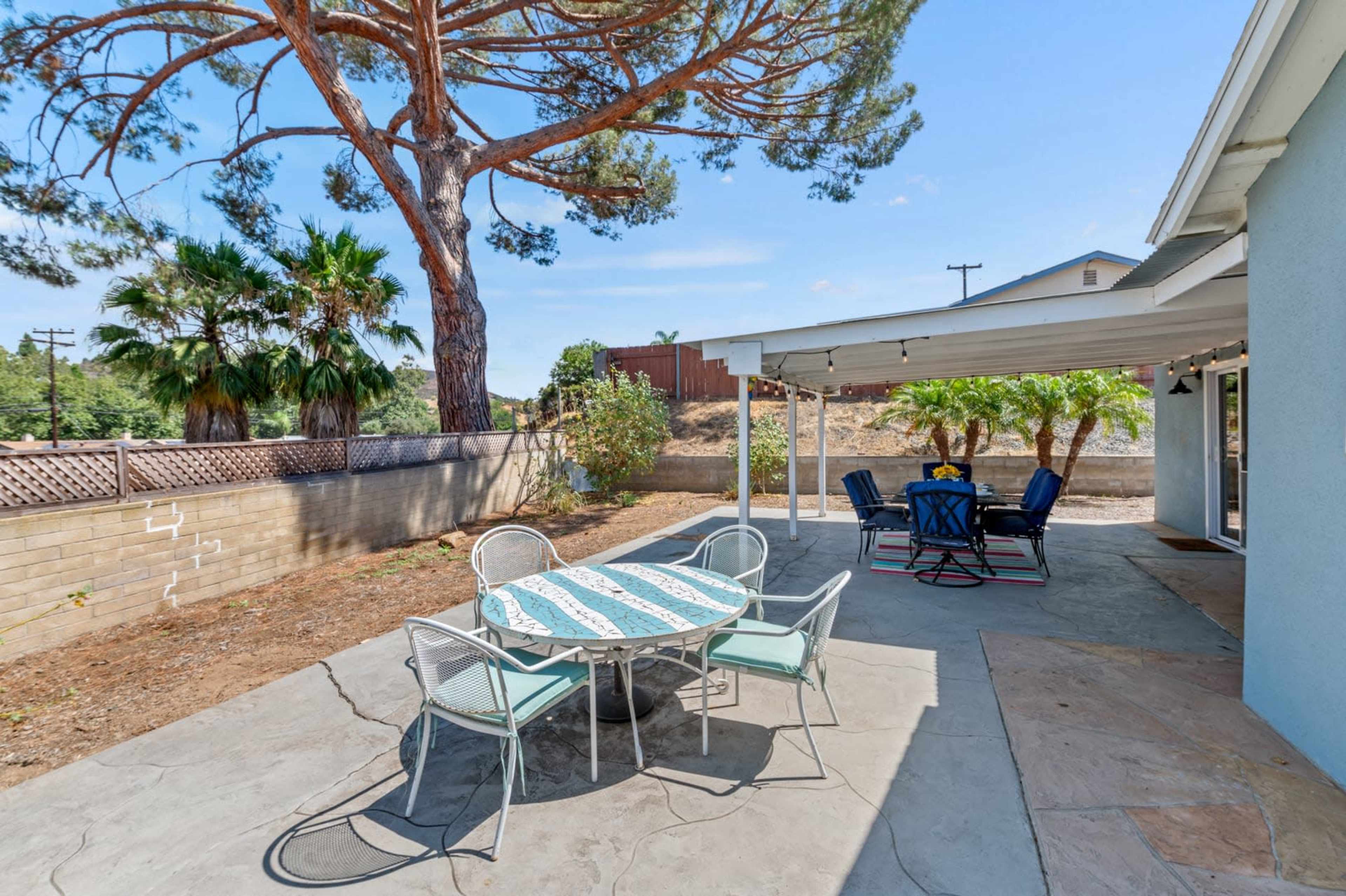 A patio area features a circular table with chairs and a shaded seating arrangement under a roof, surrounded by trees and a hillside.