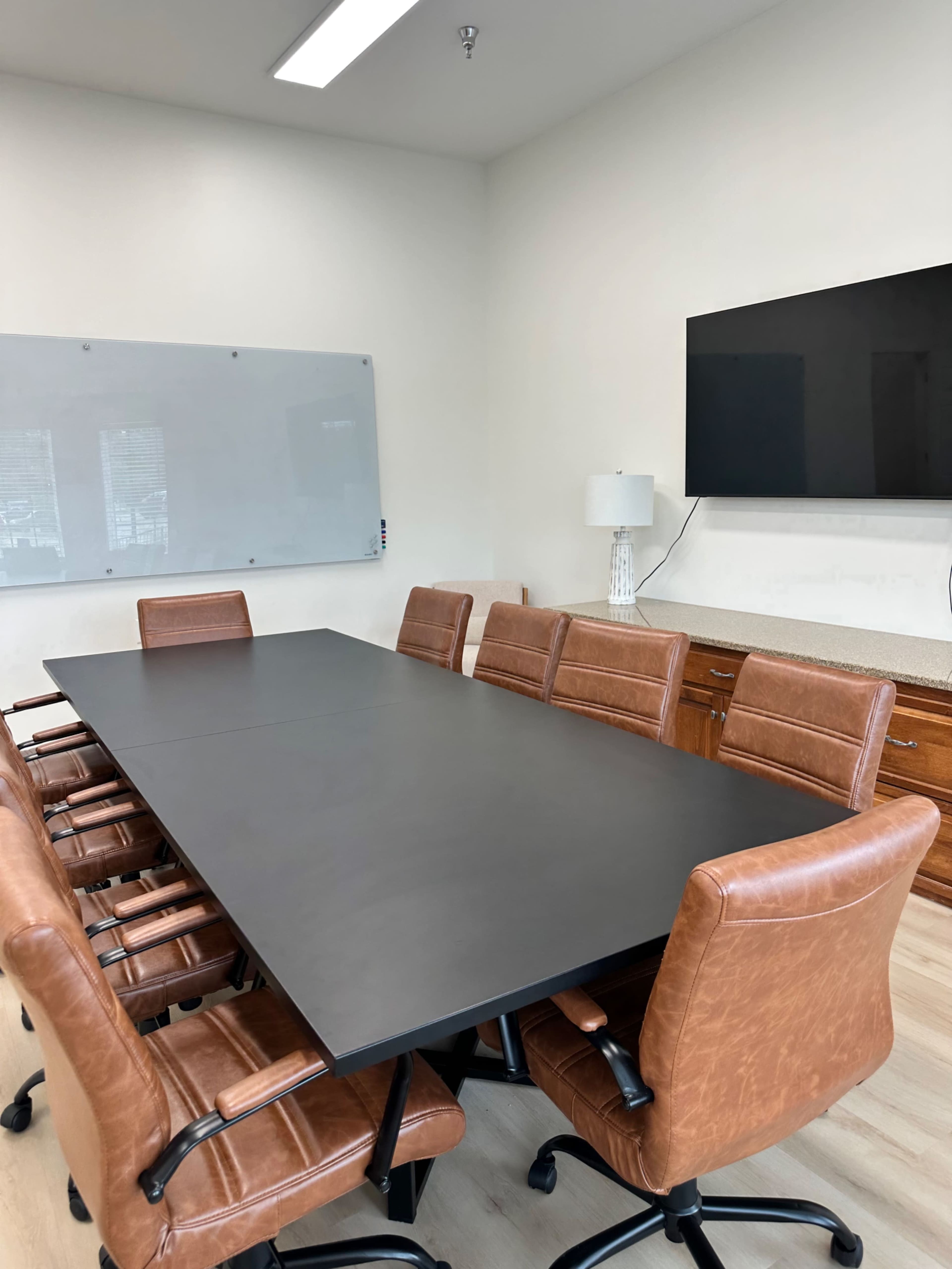 A large rectangular conference table is surrounded by brown leather chairs in a well-lit meeting room with a wall-mounted screen and a whiteboard.