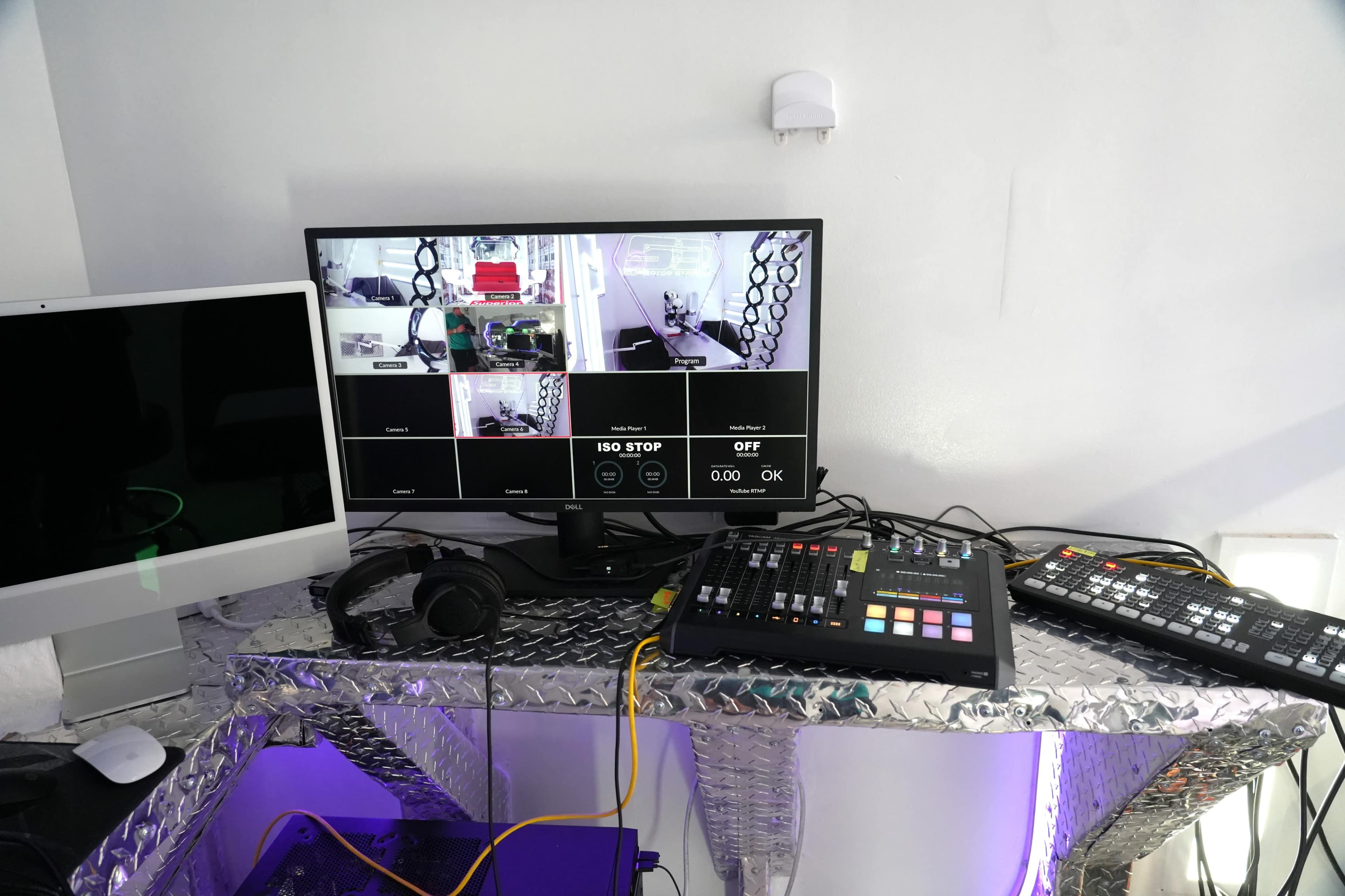 A monitor displaying multiple camera feeds is set up on a metallic desk alongside audio equipment and a computer.