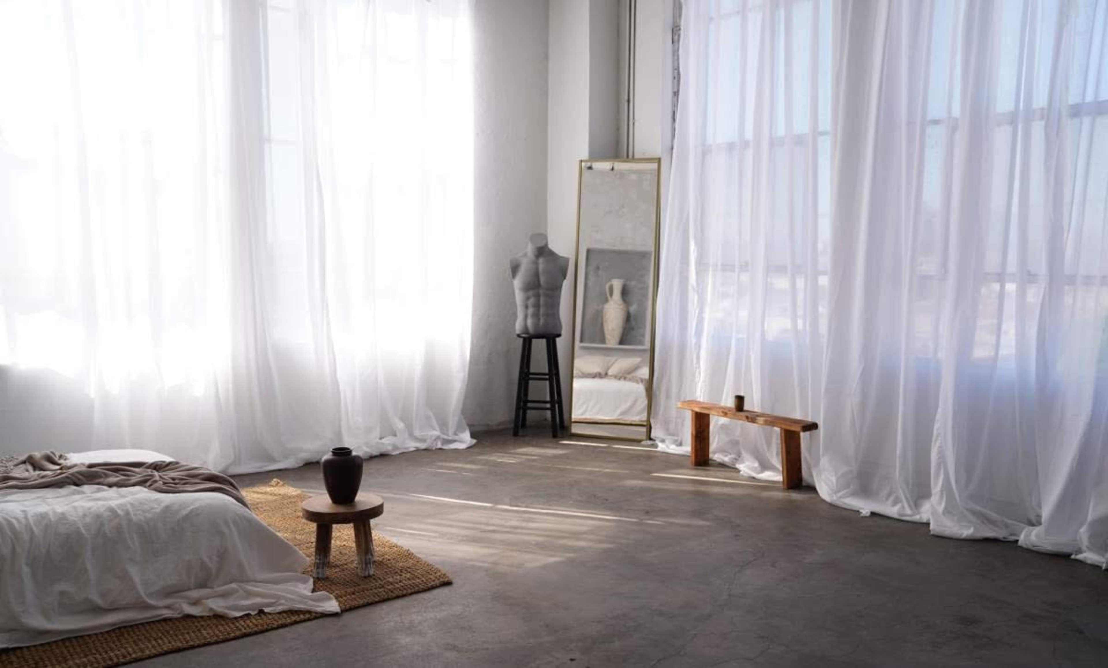 A bright, minimalist bedroom features large sheer white curtains, a bed with light bedding, a wooden stool, a pottery vase, and a full-length mirror.