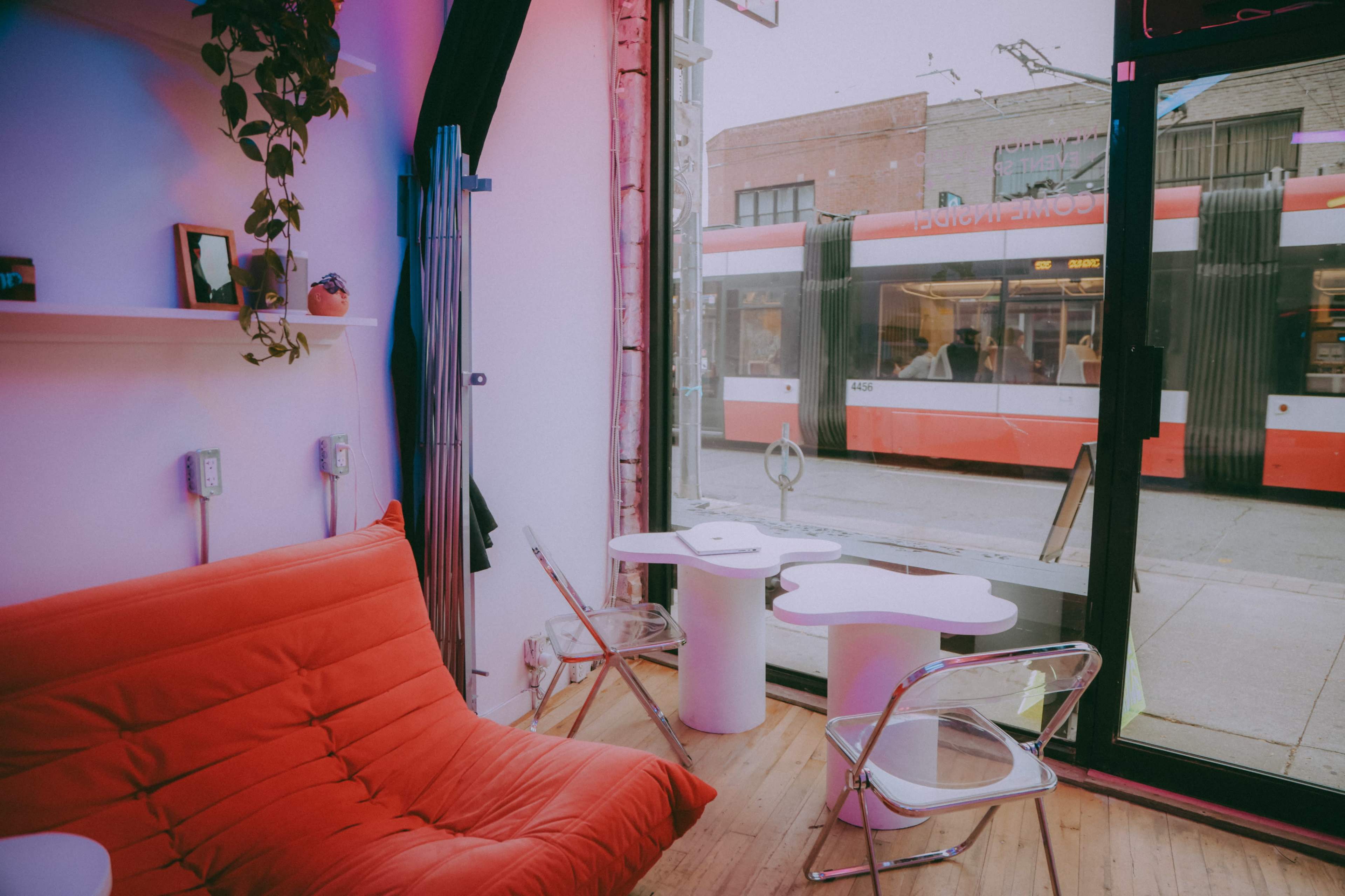 A red couch and modern chairs are arranged in a bright, minimalist interior next to a large window with a view of a tram passing by.