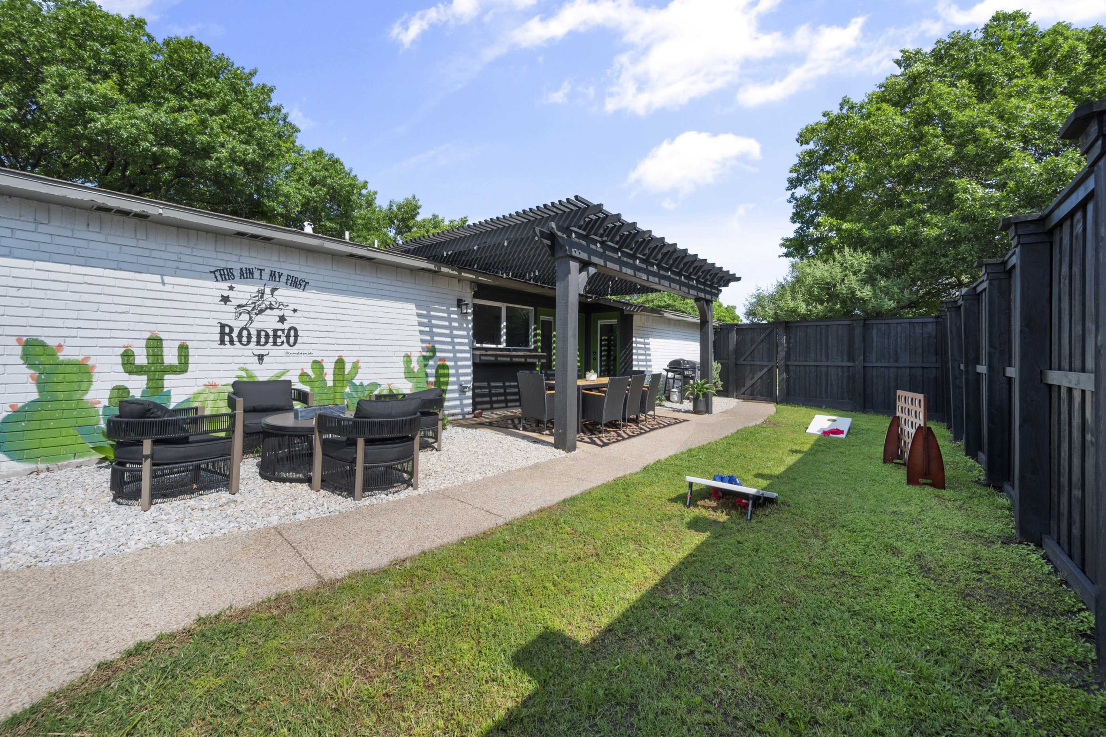 The image shows a backyard patio area with a dining table under a wooden pergola, surrounded by greenery and featuring decorative wall art of cacti.