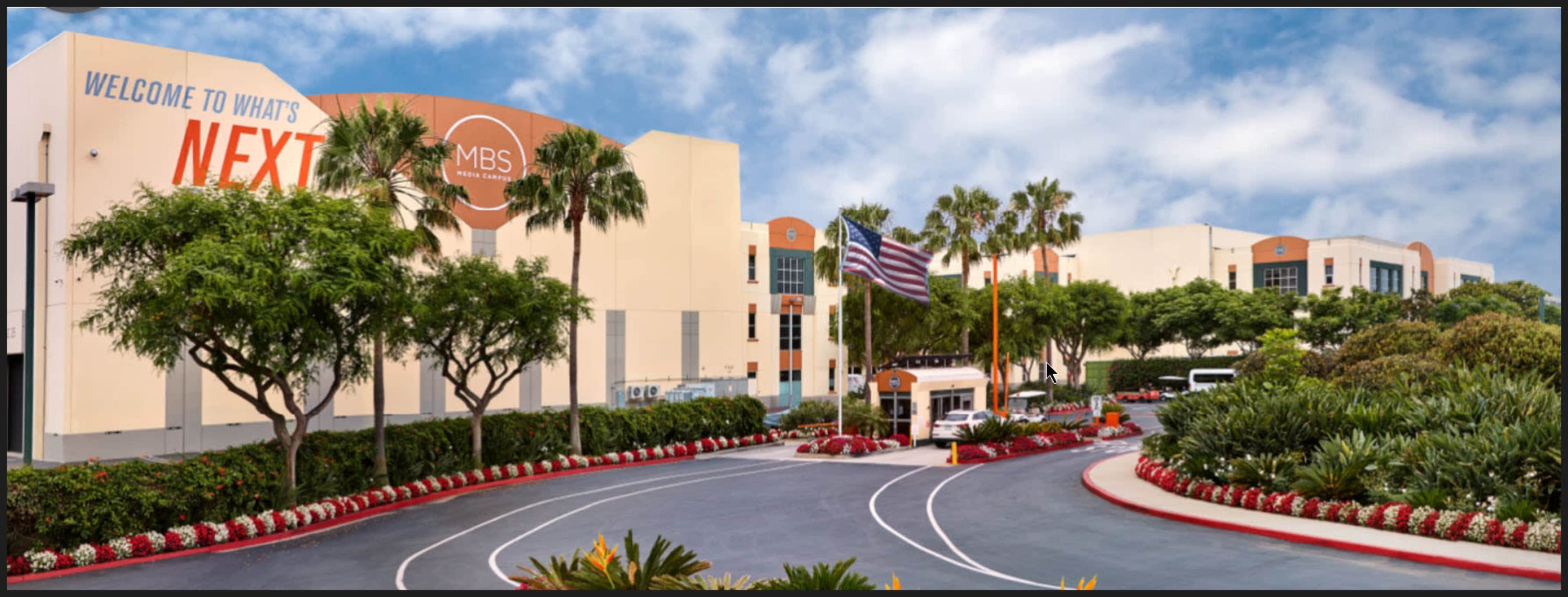 The entrance of a modern building complex with palm trees, an American flag, and a sign that reads "Welcome to What's Next."