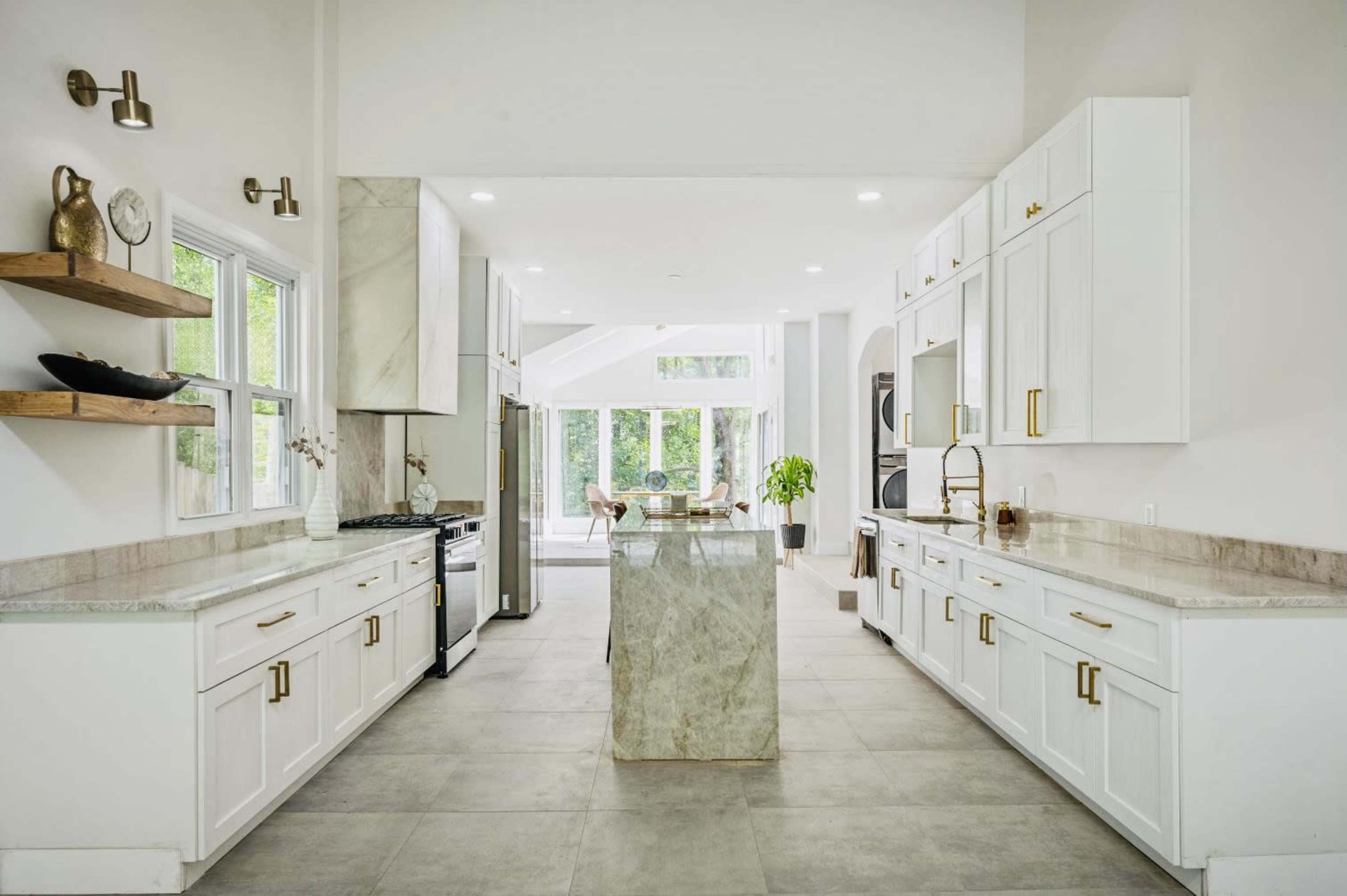 The image shows a modern kitchen with white cabinetry, a large central island, and a bright, airy layout featuring large windows that overlook a green outdoor space.