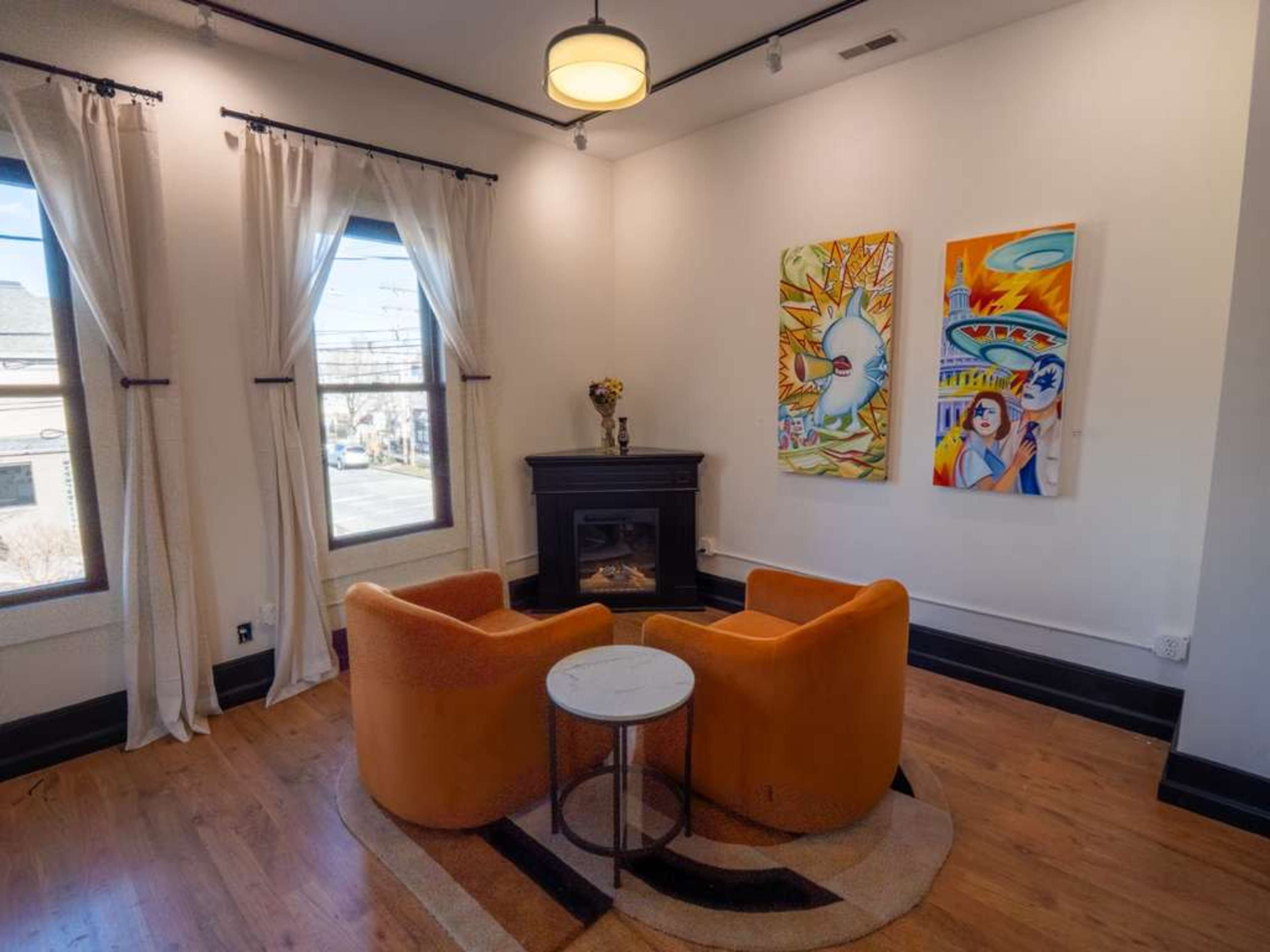 The image shows a cozy room featuring two orange armchairs, a round white table, and vibrant artwork on the walls, alongside a fireplace.