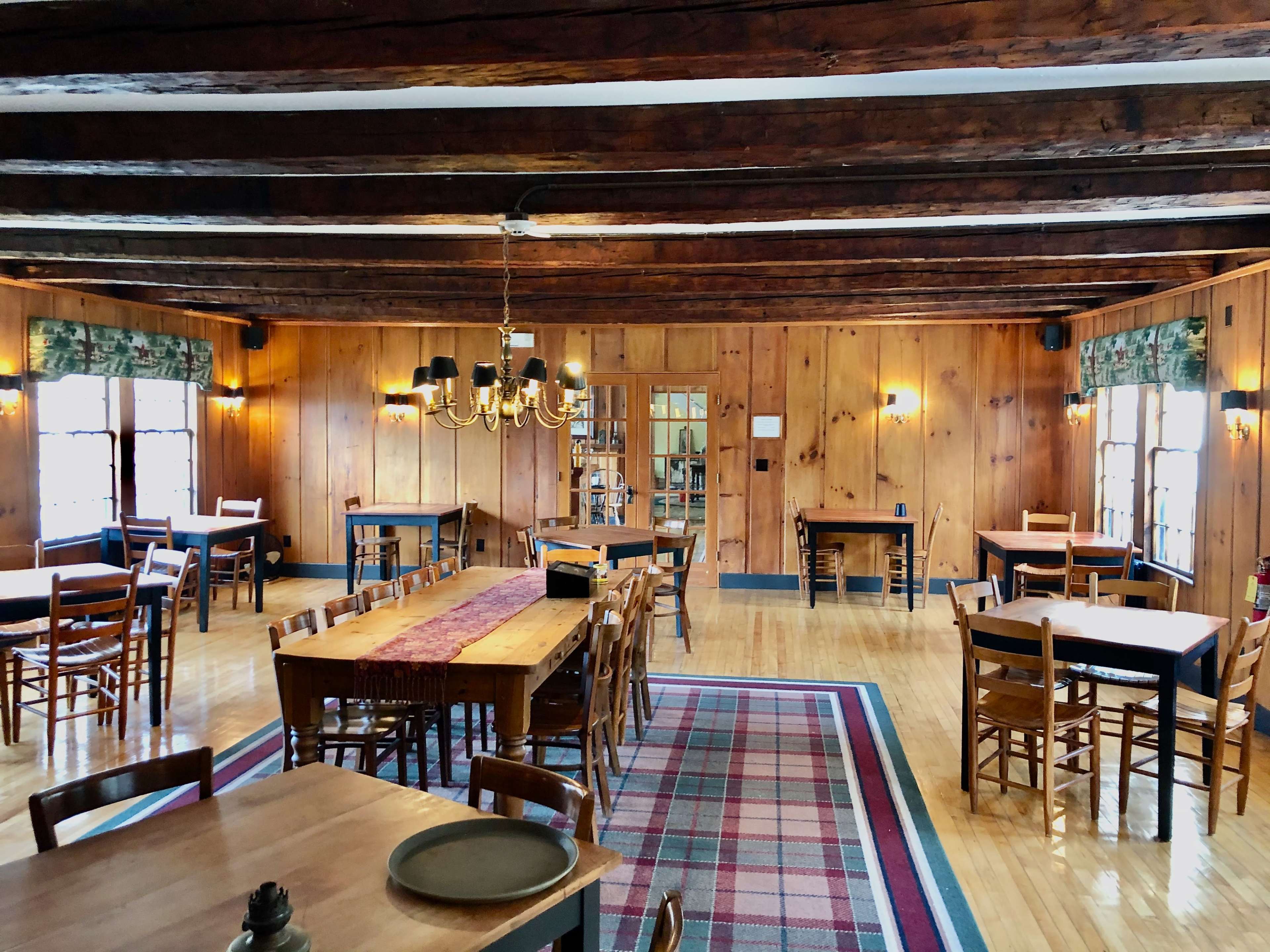 A wooden-paneled dining room with multiple tables, a chandelier, and large windows letting in natural light.