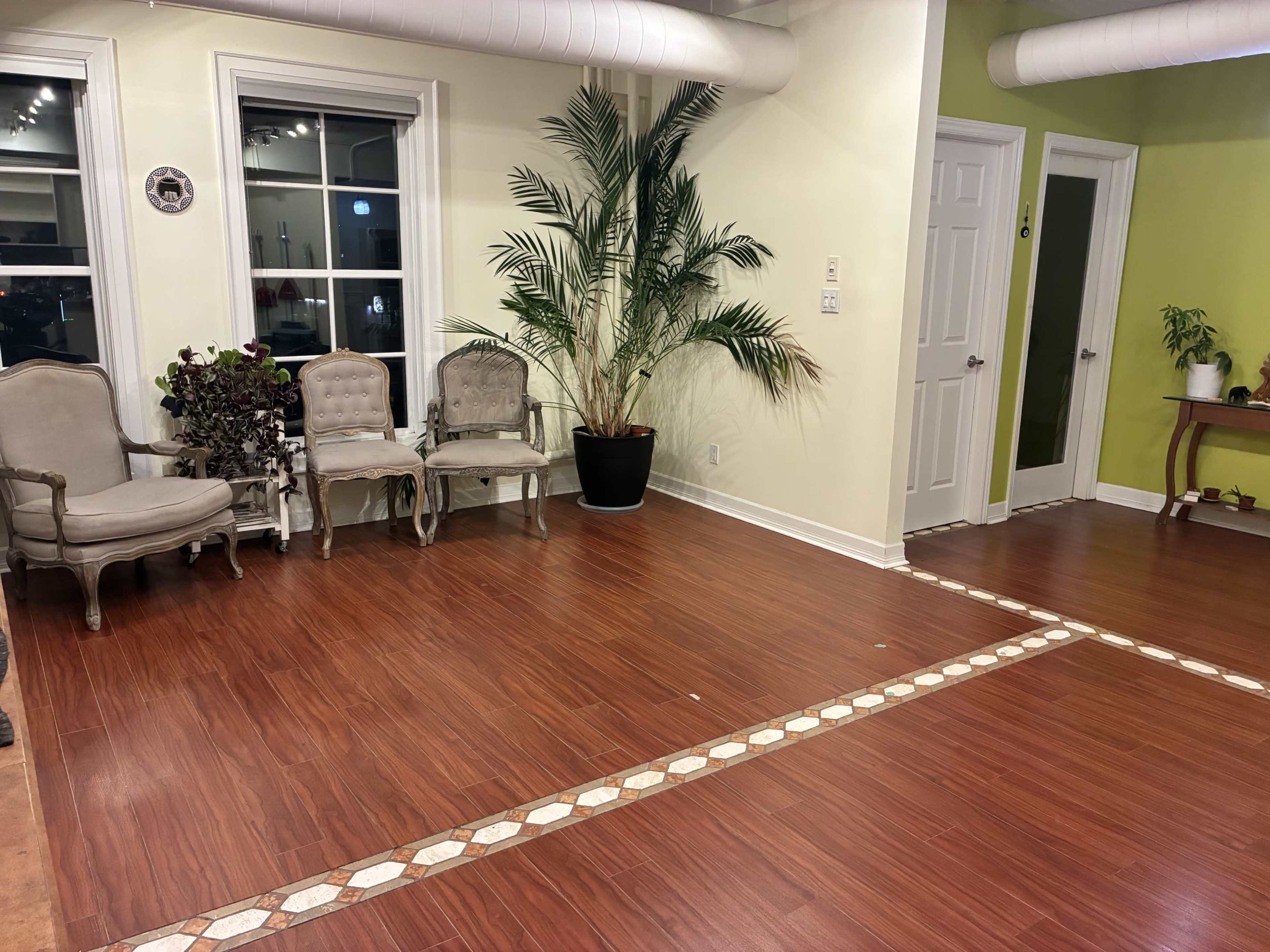 The image shows a spacious interior room with a wooden floor, a potted palm plant, two upholstered chairs, and a small table against a green wall.