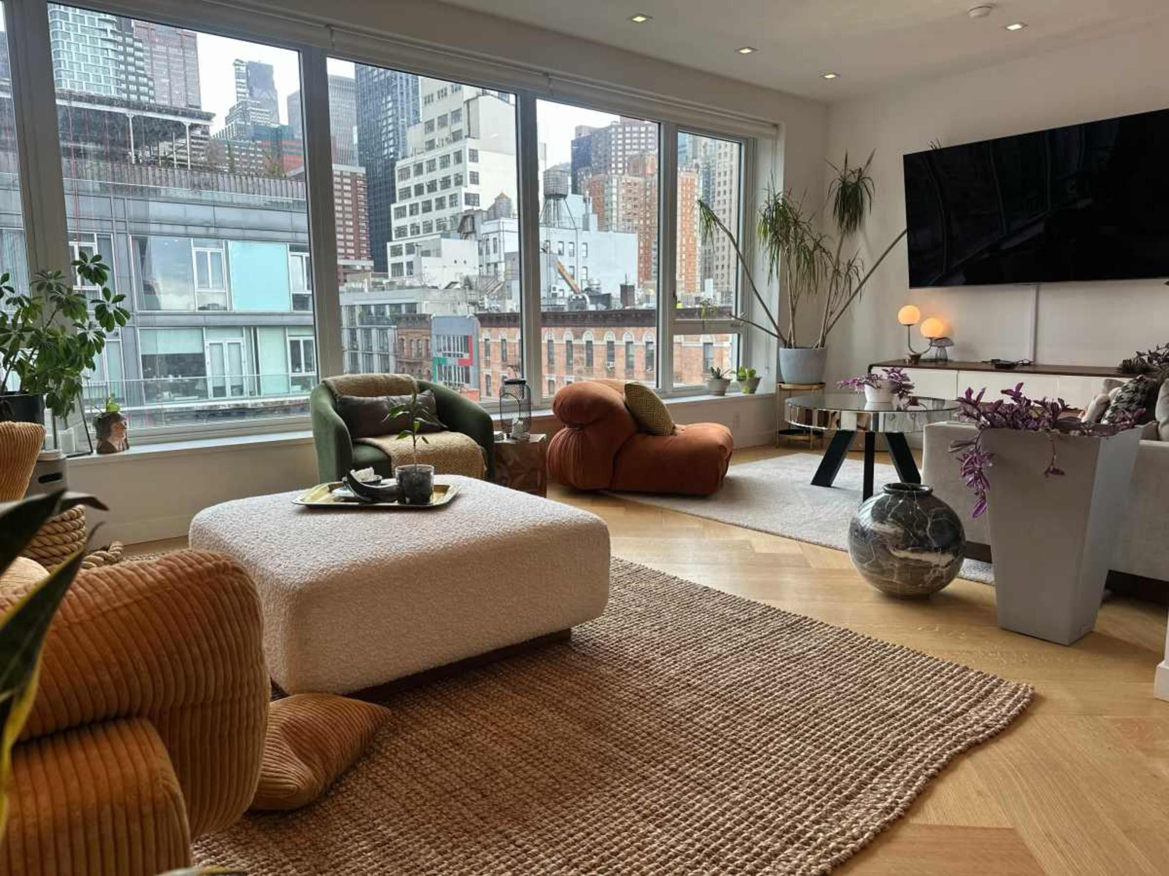 The image shows a modern living room with a large window overlooking a cityscape, featuring contemporary furniture and decorative plants.