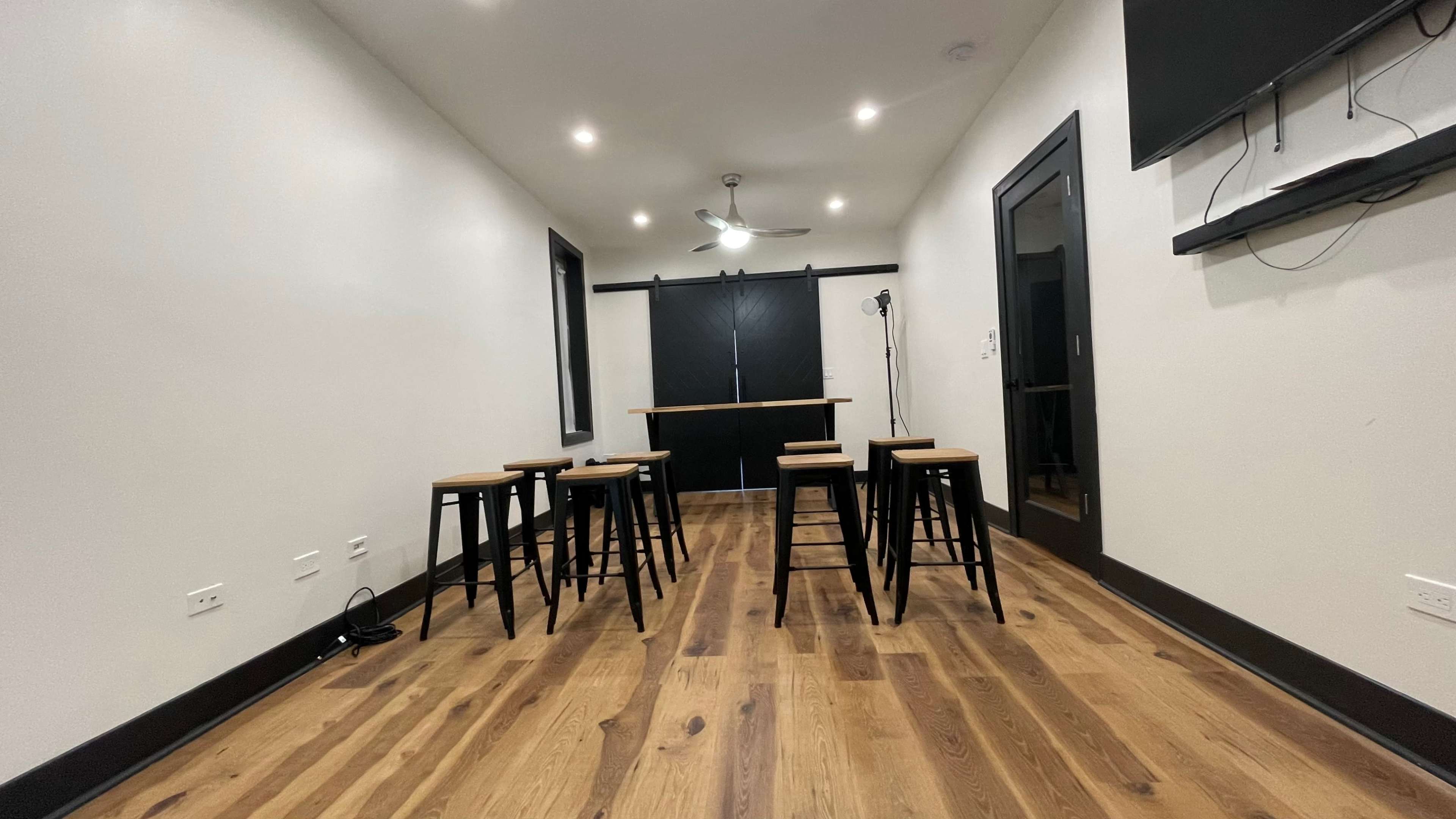 The image shows a room with wooden flooring, featuring several tall black stools positioned around a central area, a ceiling fan, and a large black sliding door at the far end.