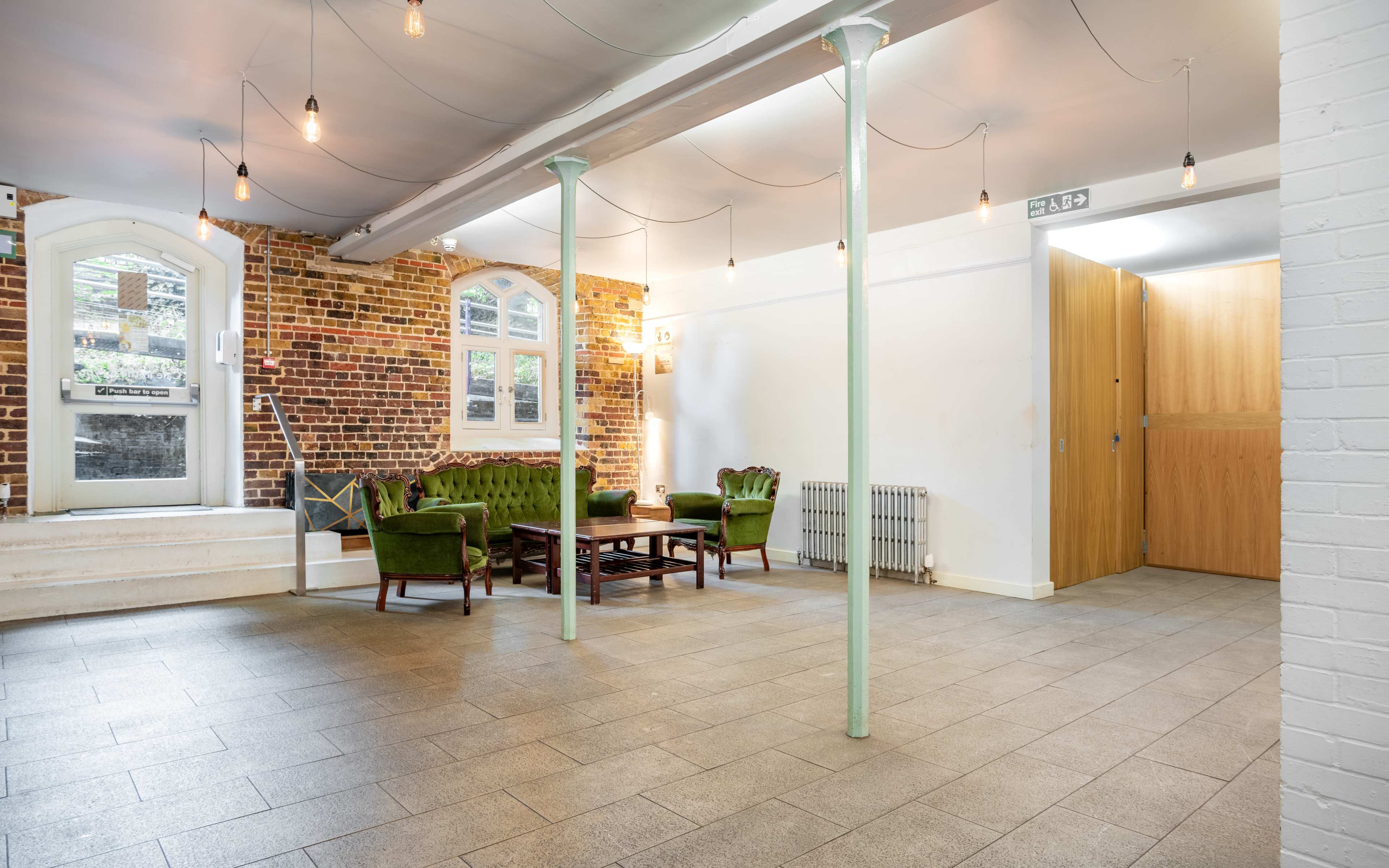 An interior space with a green seating area, a wooden table, and a doorway leading to another room, all against a backdrop of exposed brick walls and hanging light bulbs.