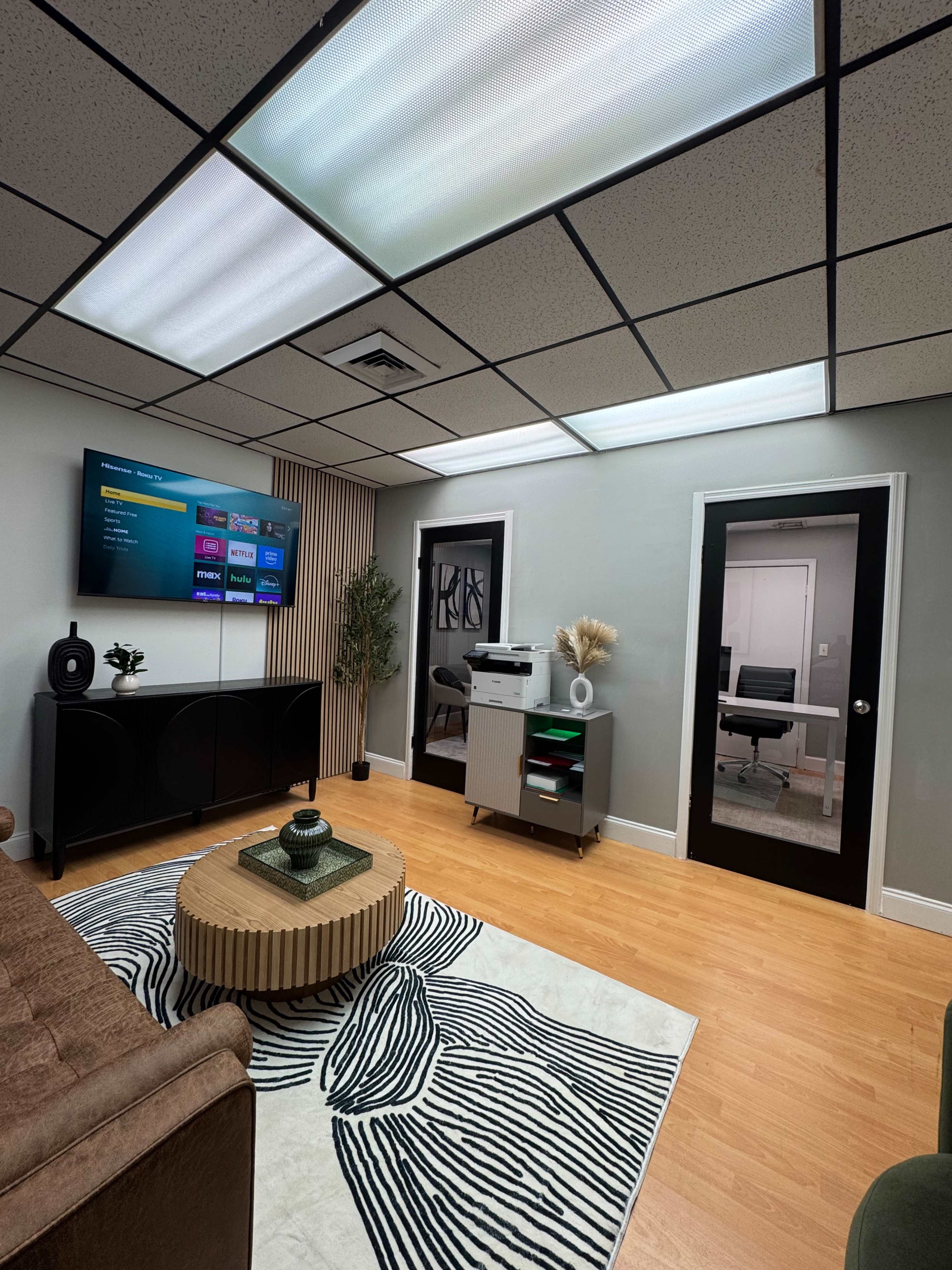 The image depicts a modern office space featuring a seating area with a brown sofa, a patterned area rug, a television mounted on the wall, and a door leading to another room.