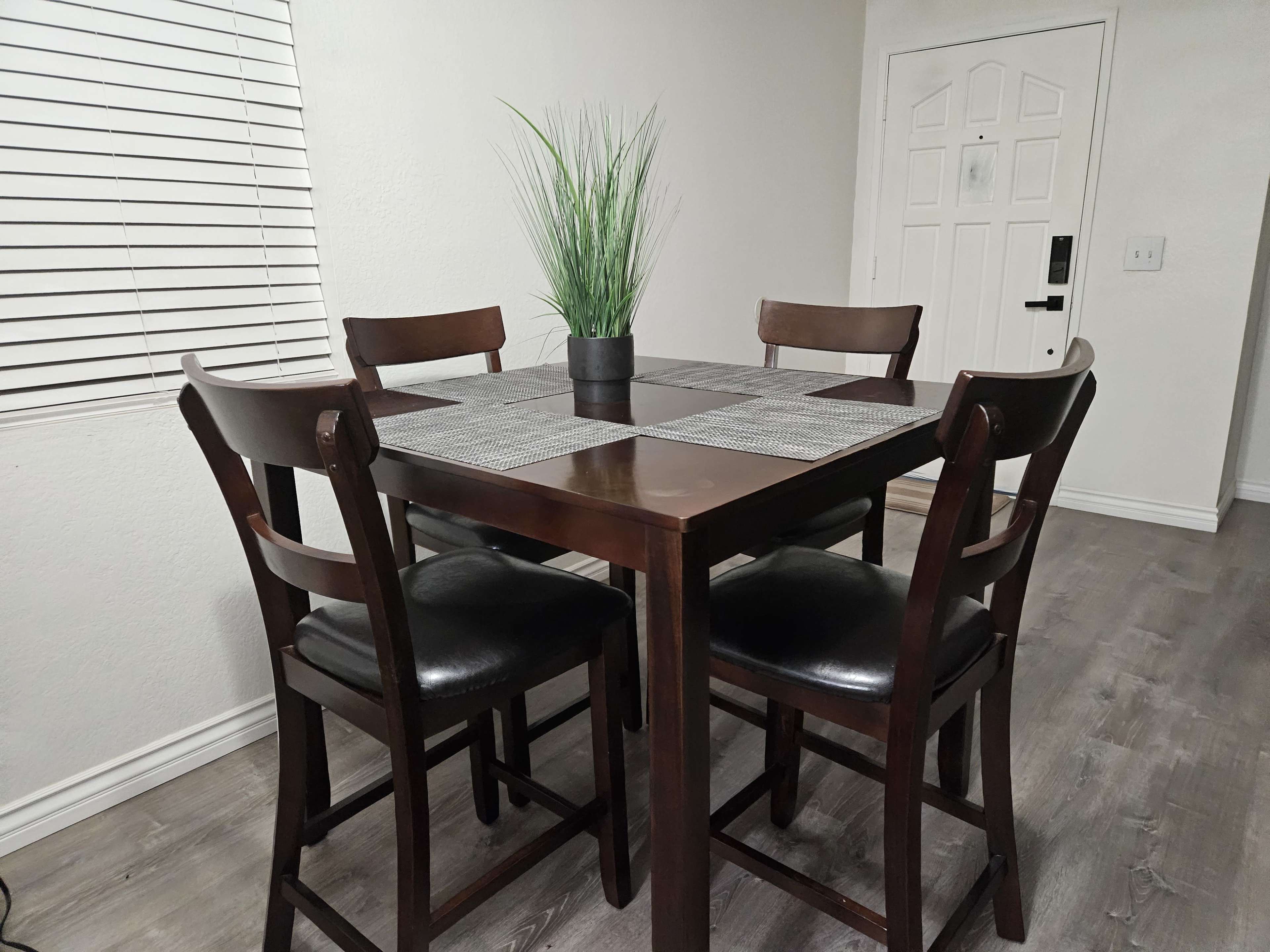 A dining table with four high-backed chairs and a small potted plant centerpiece is set against a light-colored wall with a door and window nearby.