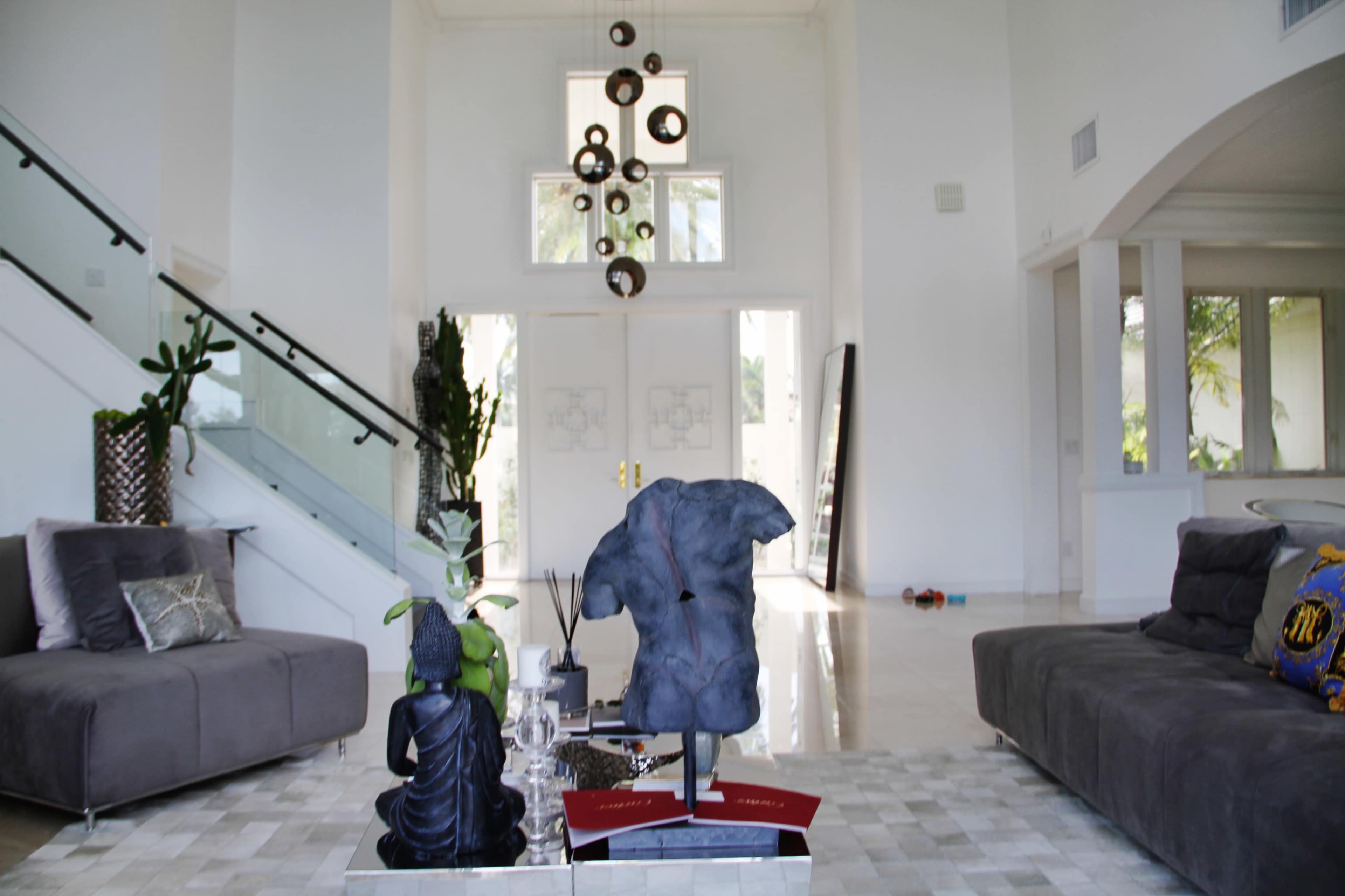 A spacious modern living room with a high ceiling, featuring a sculpture in the center, a gray sofa on either side, and large glass doors at the back.