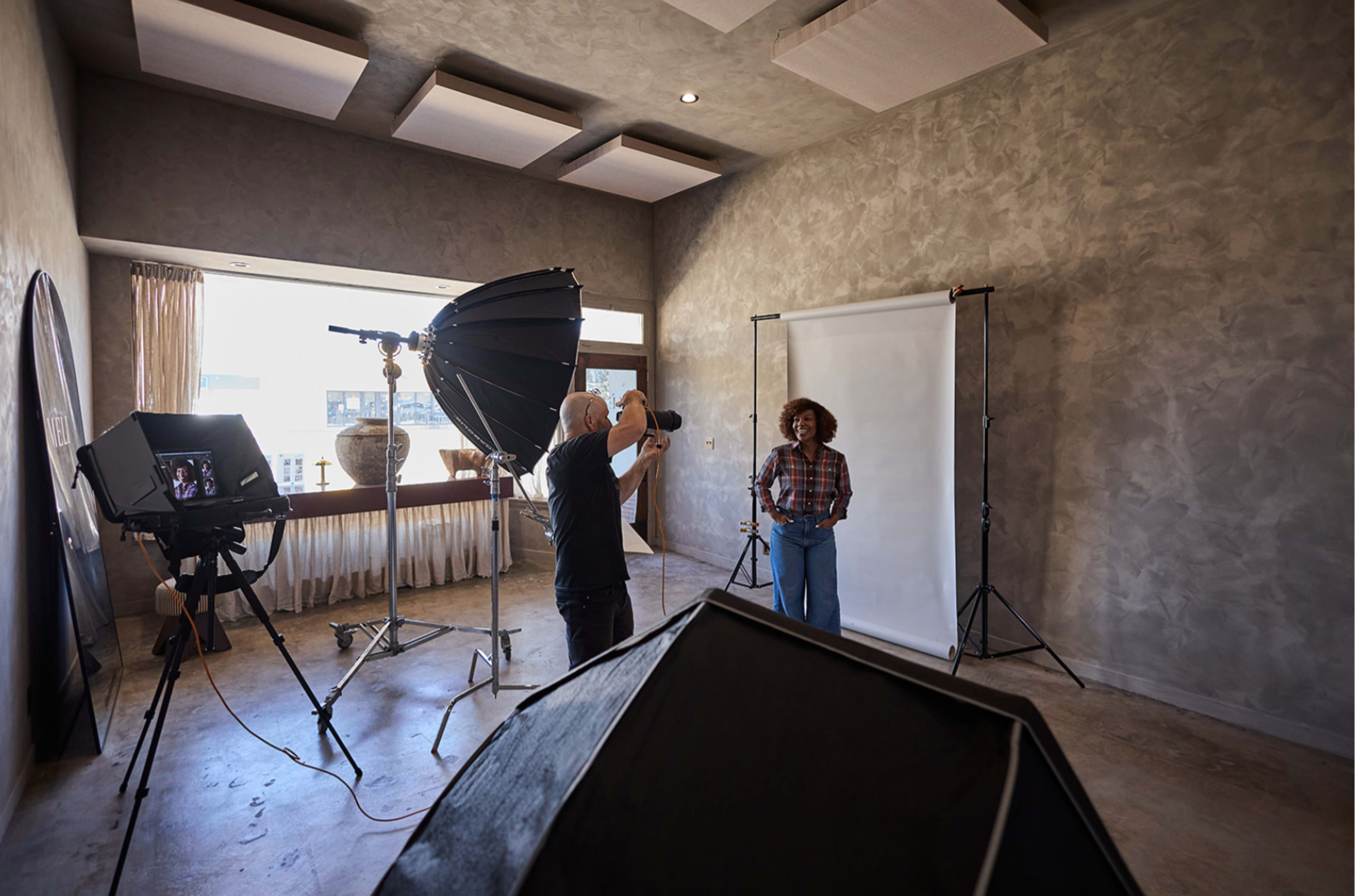 A photographer is capturing an image of a woman posing in a studio filled with soft lighting and backdrops.