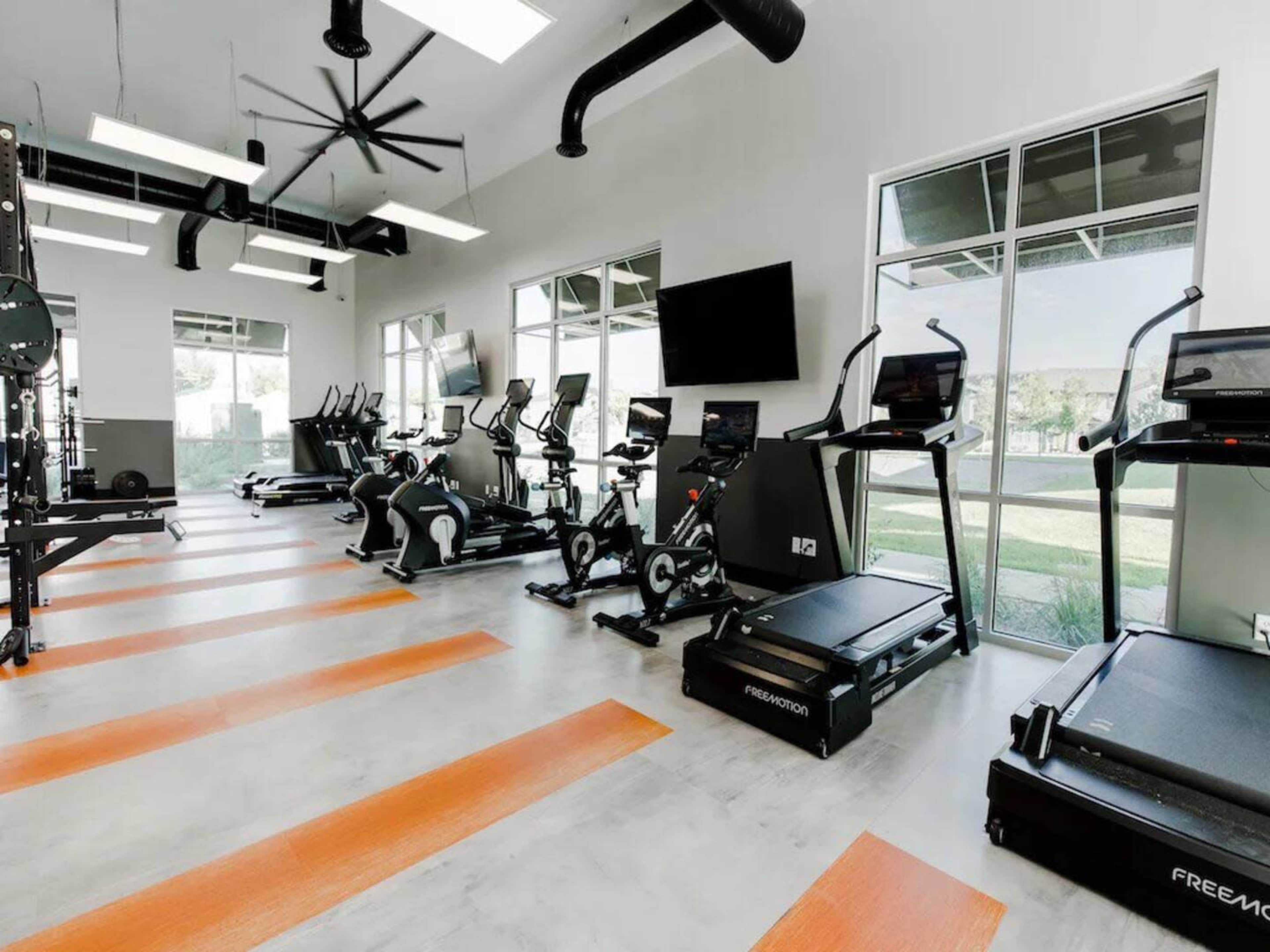 The gym features several exercise machines, including treadmills and stationary bikes, arranged neatly in a well-lit space with large windows.
