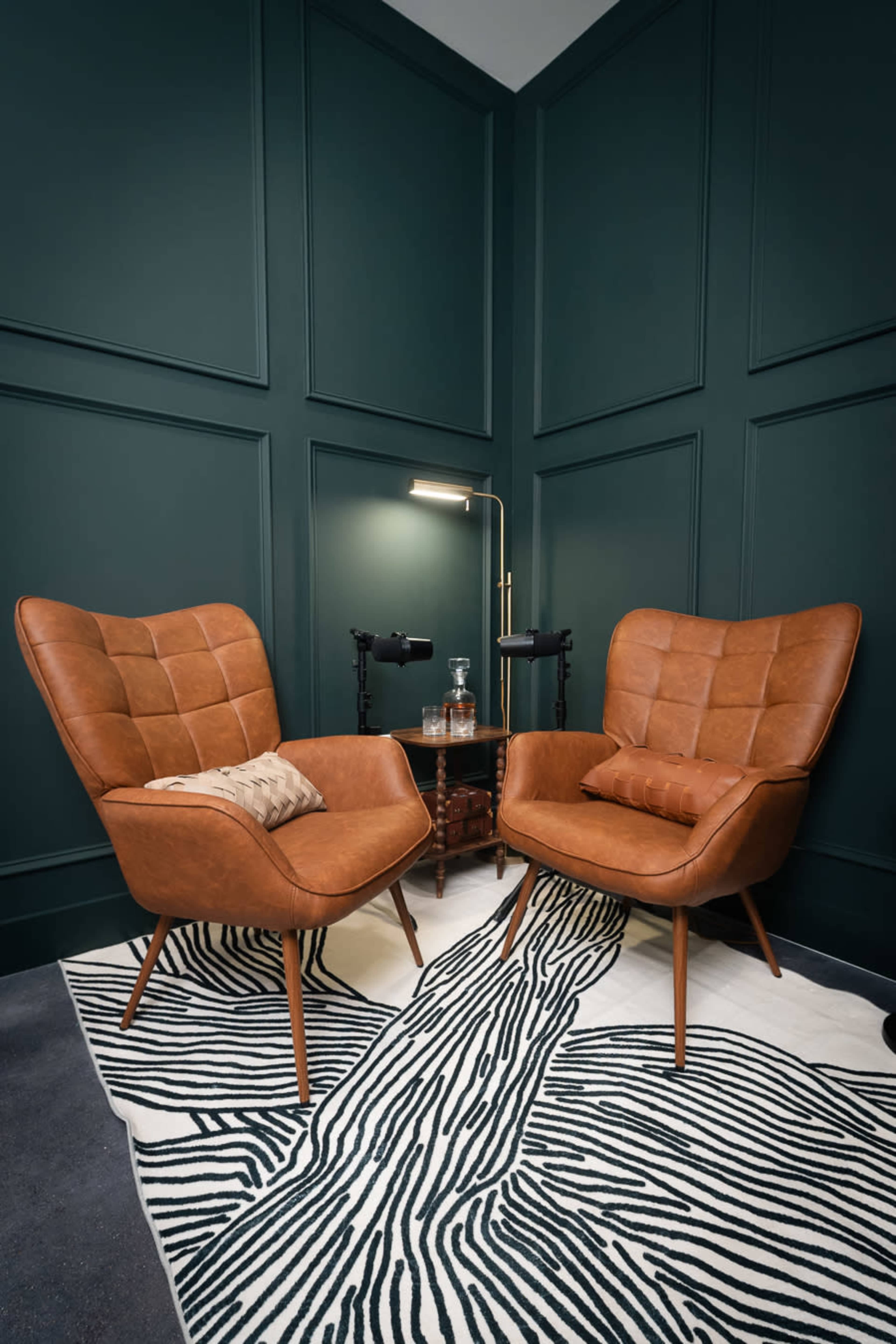 The image shows two brown leather chairs arranged closely together on a zebra-patterned rug, next to a small table with a lamp and water carafe, against a dark green paneled wall.