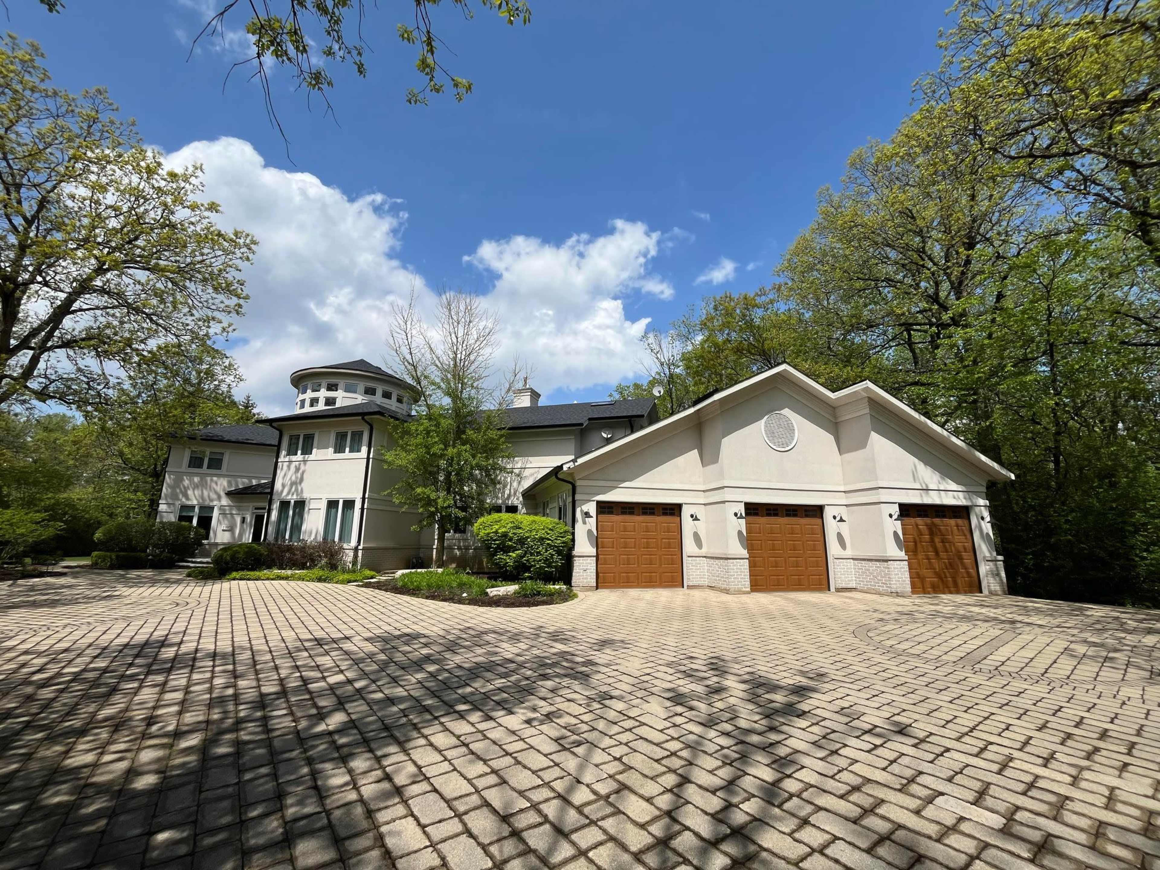 A large, modern house with a circular turret and a three-car garage is set among trees under a partly cloudy sky.
