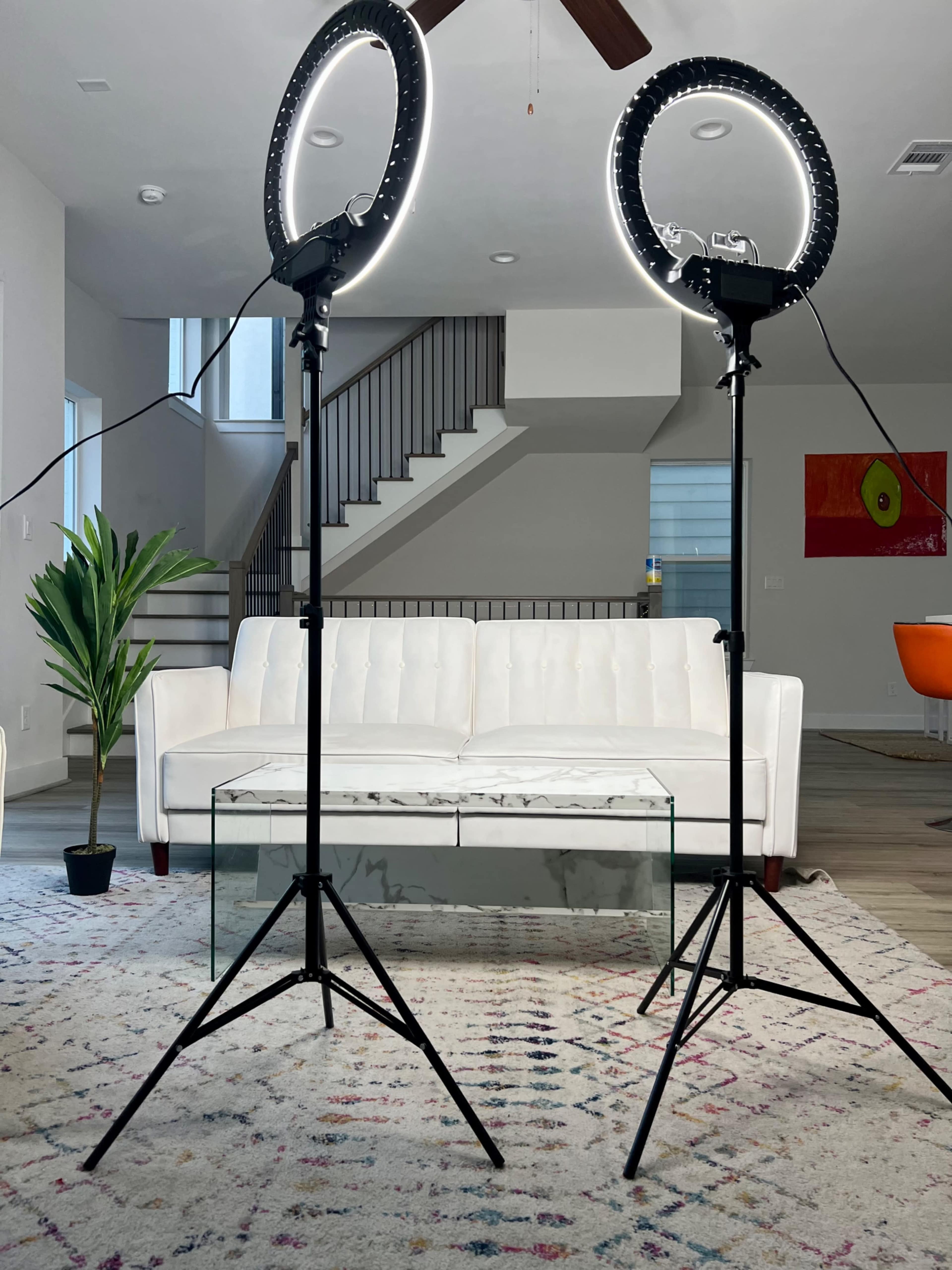 The image shows two ring lights on stands facing a white sofa with a glass coffee table, in a modern living room setting with a staircase in the background.
