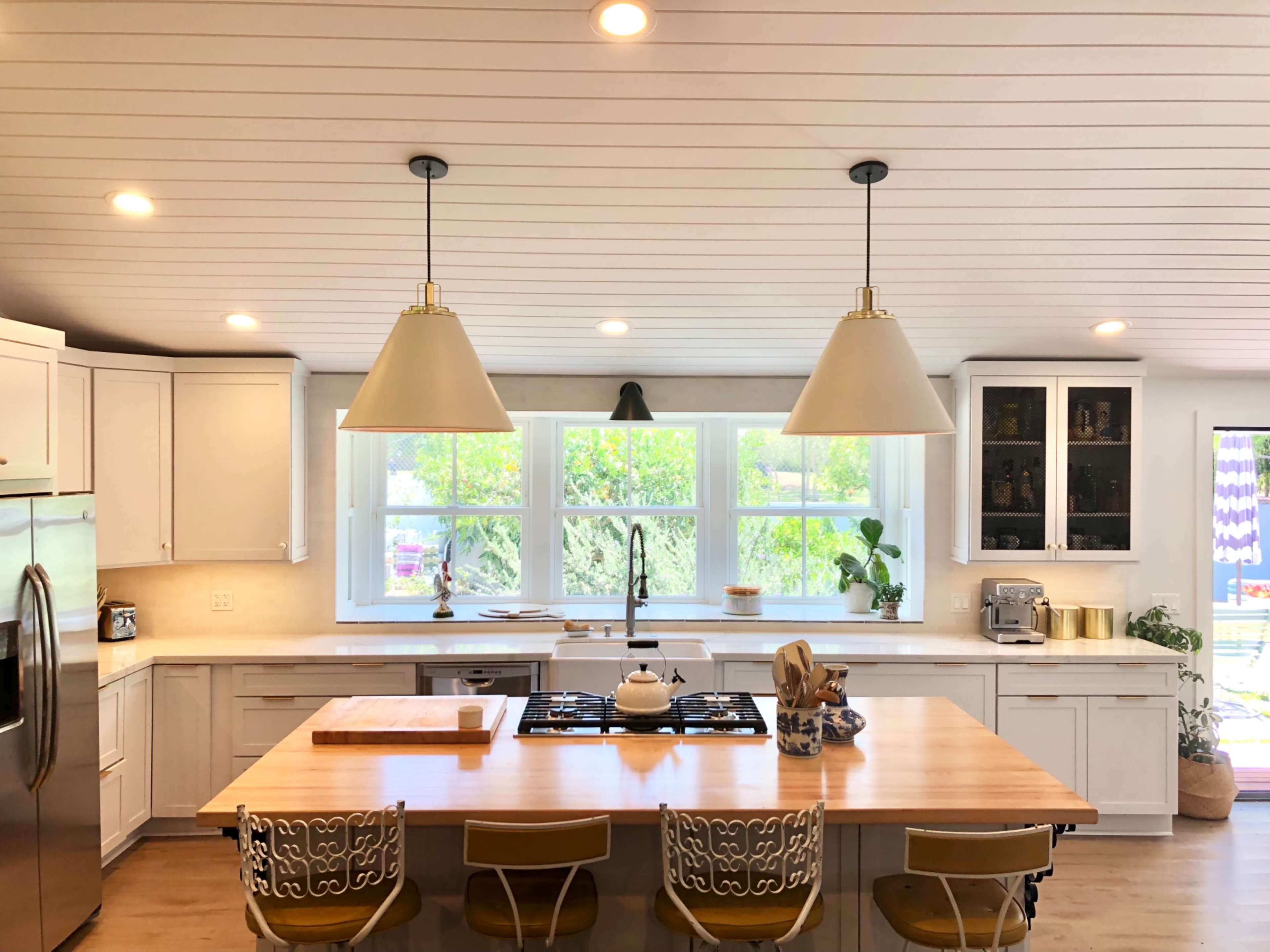 A modern kitchen featuring a central island with a cooktop, pendant lights, and white cabinetry, illuminated by natural light from large windows.