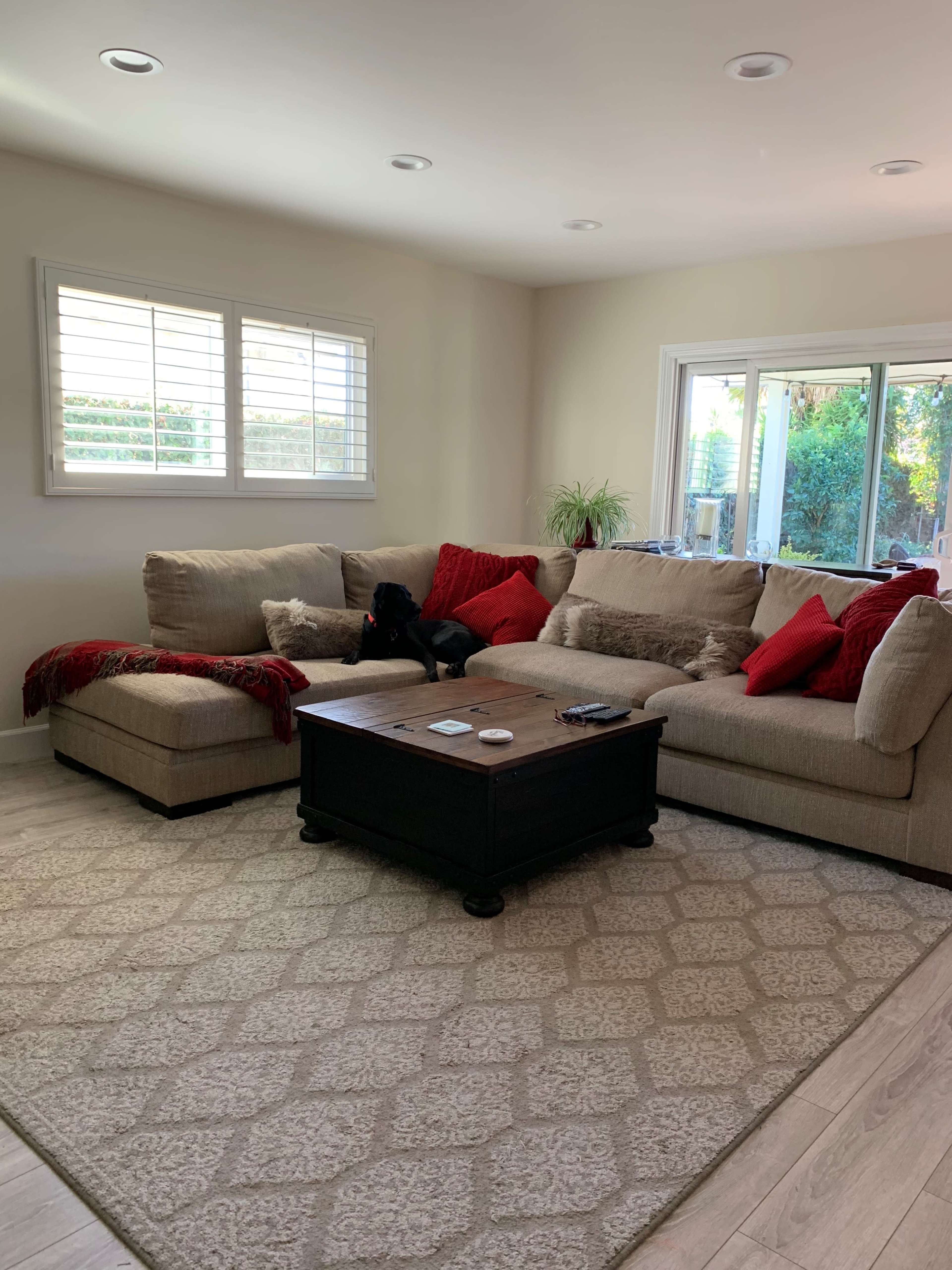 A spacious living room features a large beige sectional sofa with red accent pillows, a dark wooden coffee table, and large windows overlooking greenery.