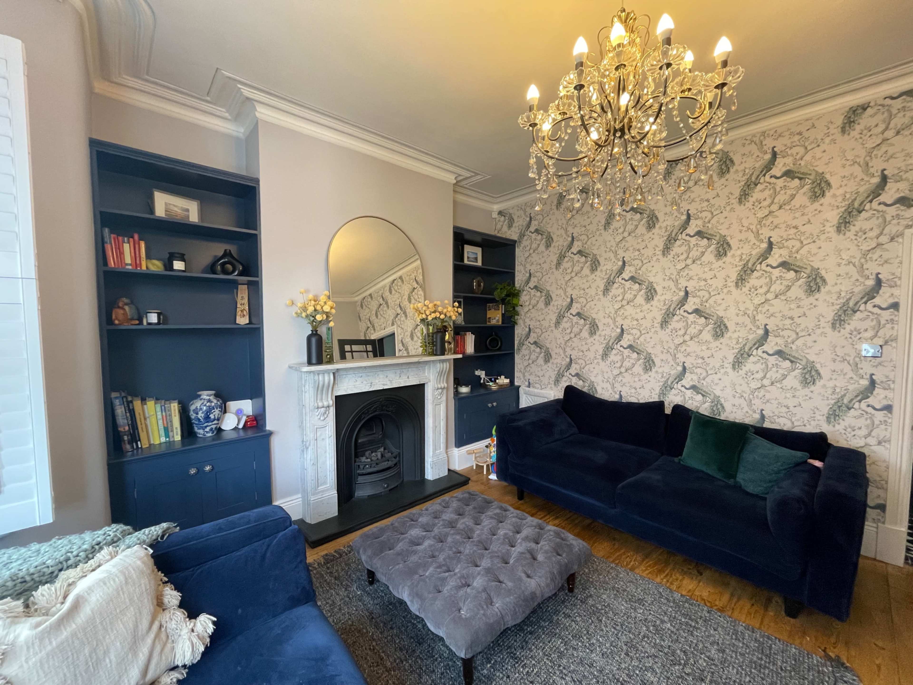 The living room features a fireplace, a chandelier, and dark blue bookshelves adorned with decorative items against a patterned wallpaper backdrop.