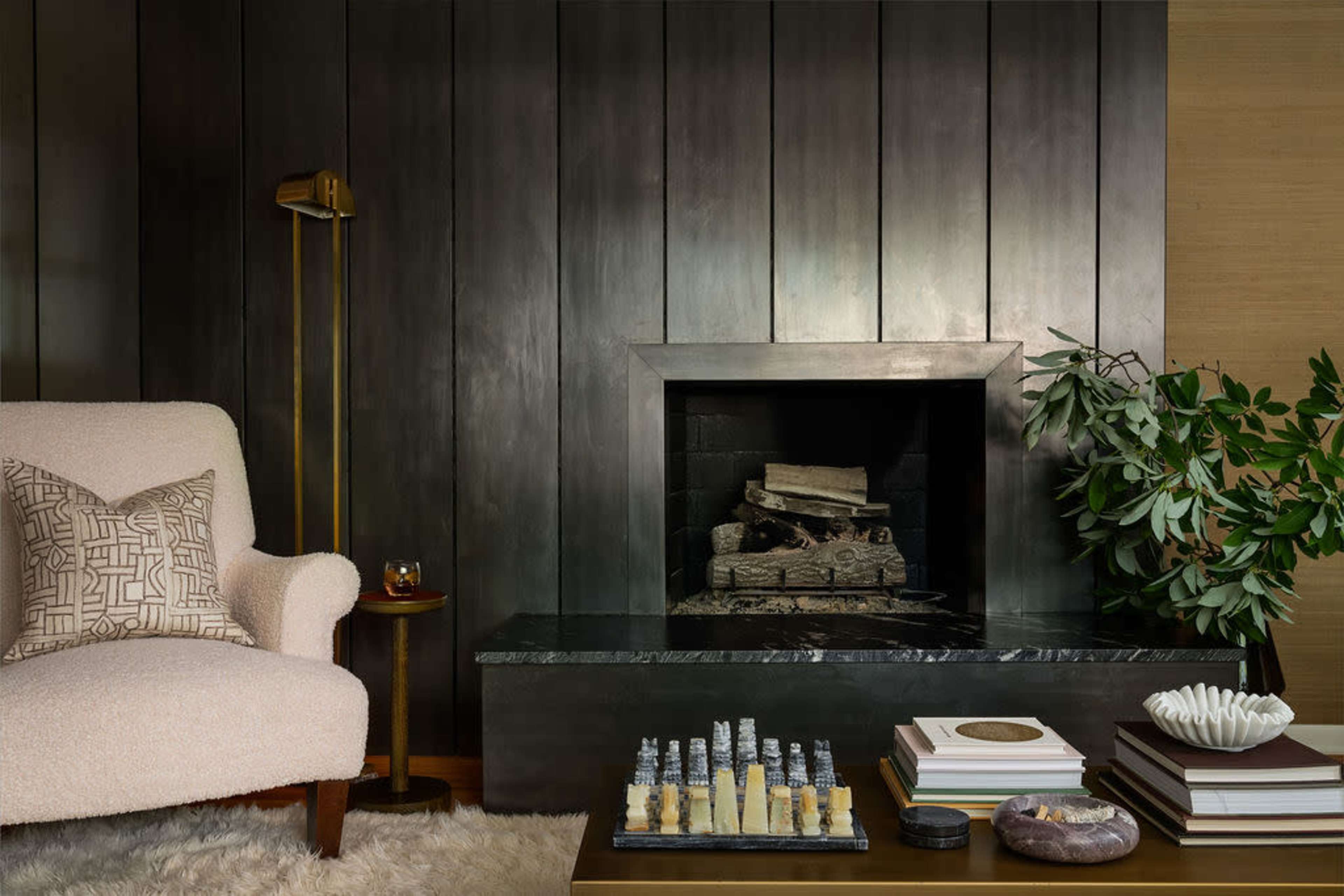 A cozy living room features a beige armchair and a sleek fireplace with stacked firewood, surrounded by decorative items on a coffee table.