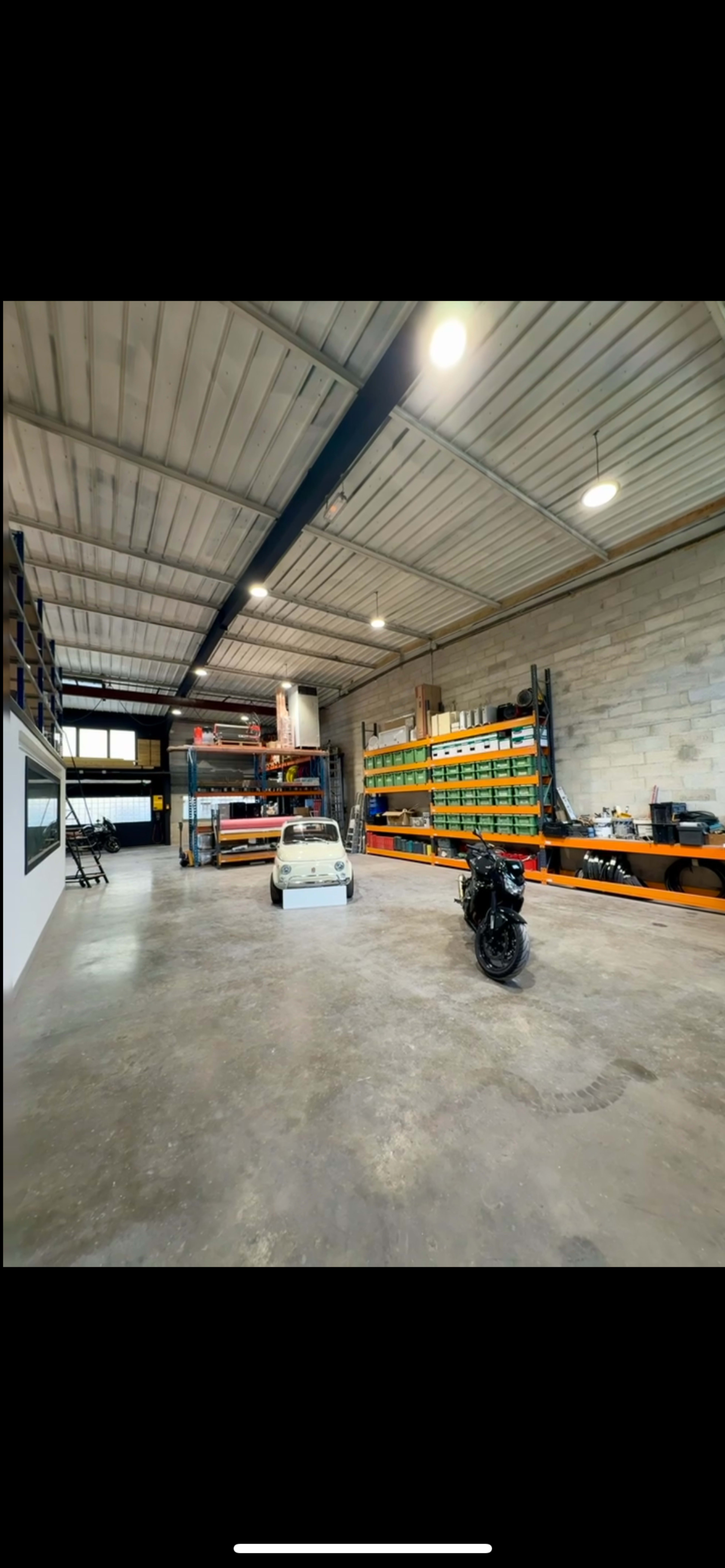 The image shows a spacious garage with concrete flooring, shelves stocked with various items on the walls, and a small white vehicle alongside a motorcycle in the center.