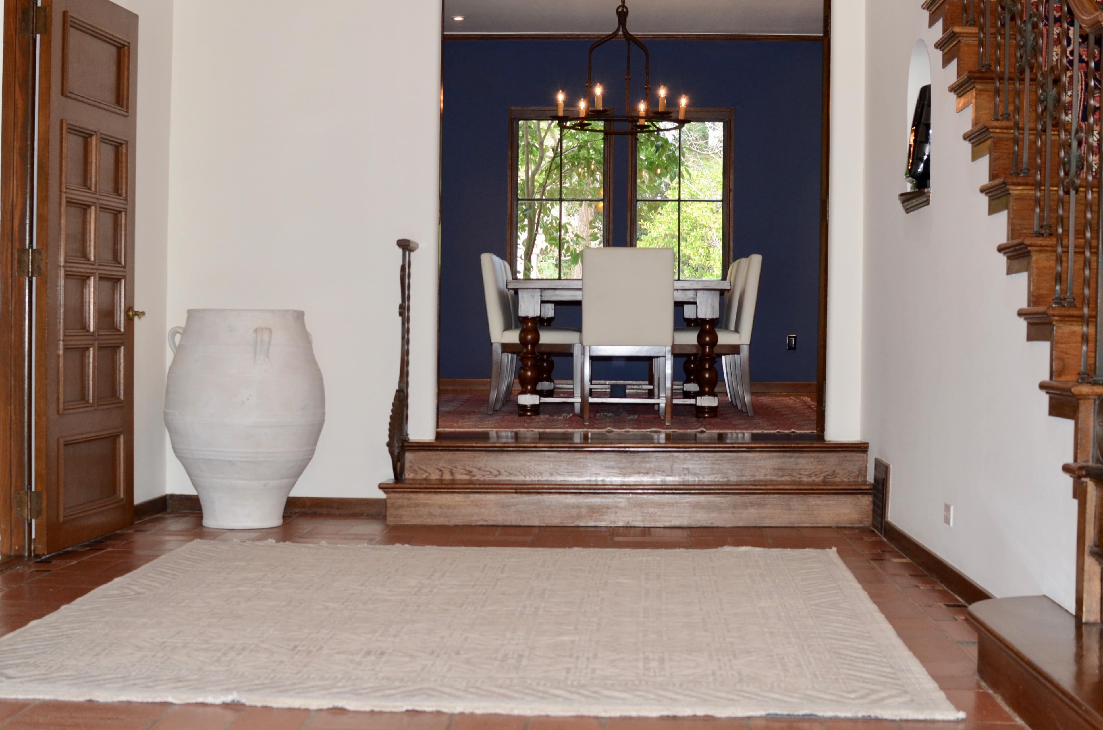 The image shows a spacious foyer with a large white vase and a beige rug that leads to a dining area with a blue wall and a wooden table set for six.