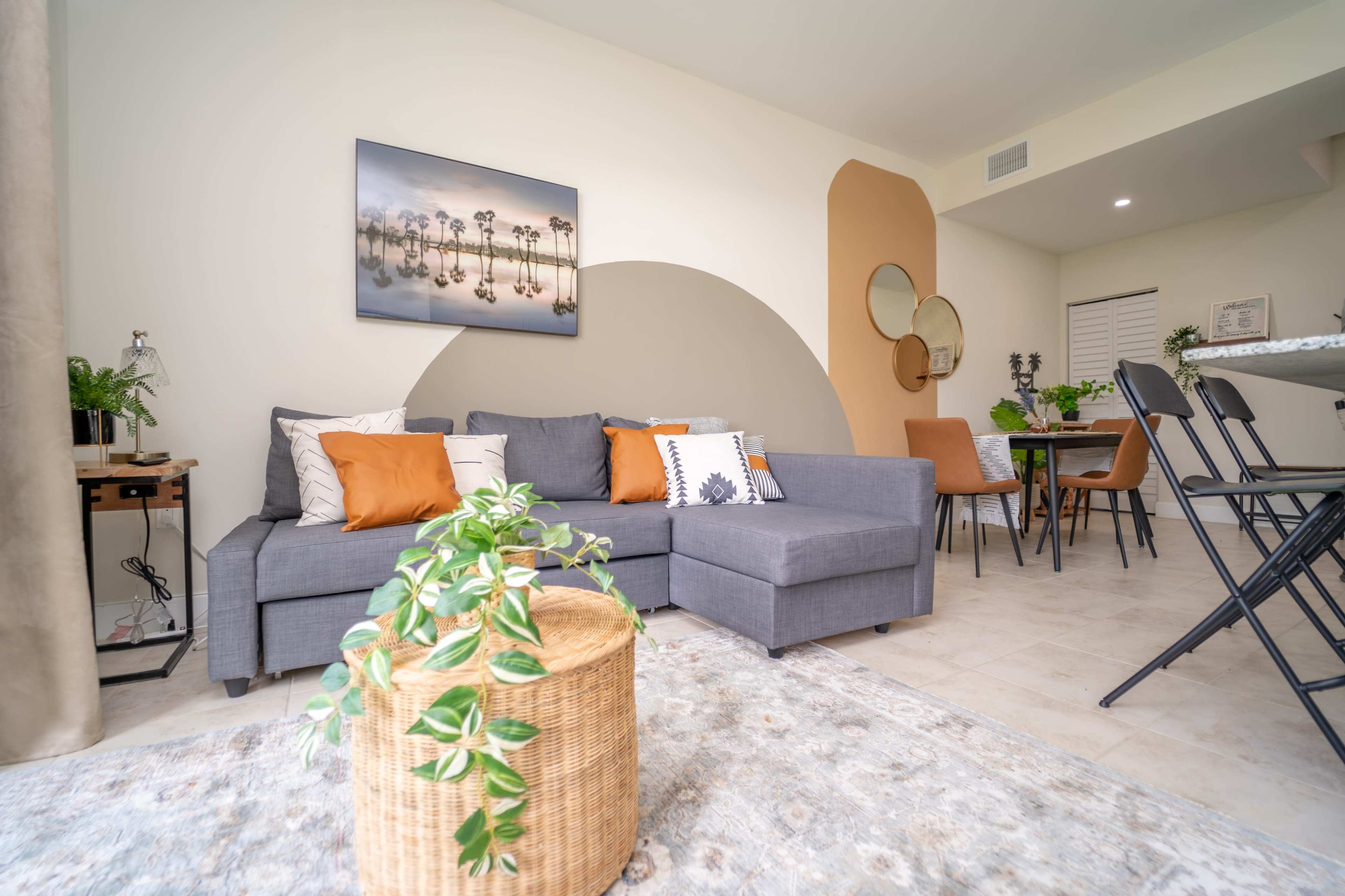 The image shows a modern living room with a gray sectional sofa, a round woven coffee table, and a dining area featuring a table and chairs.