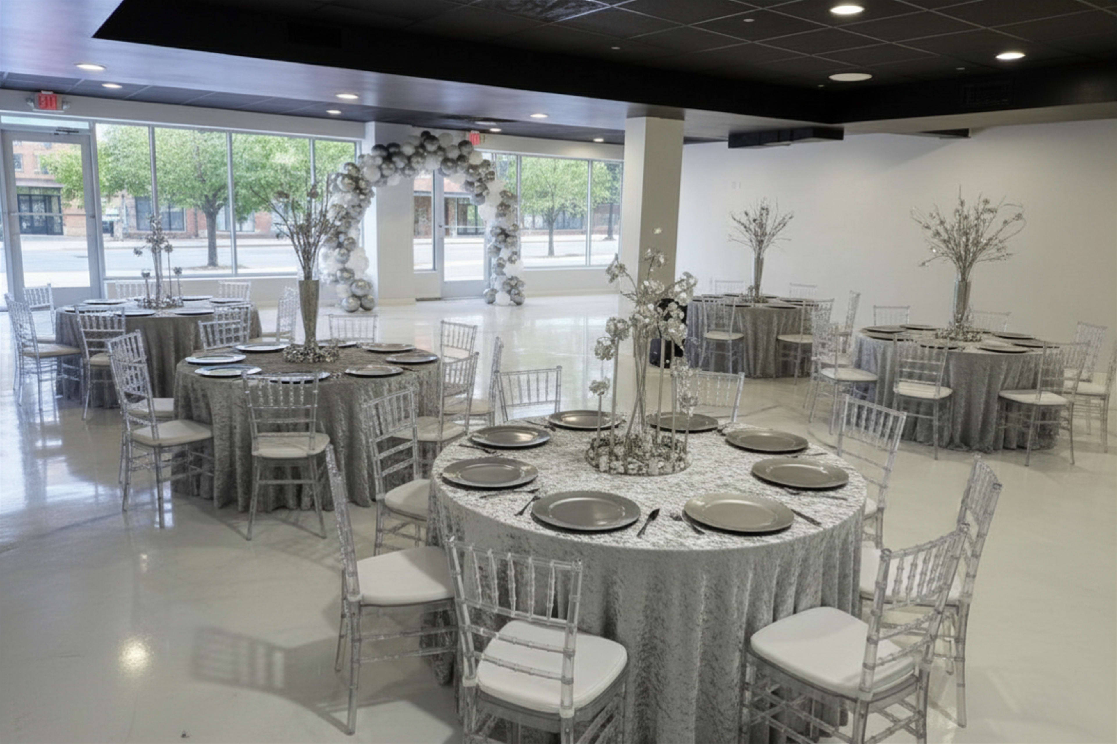 The image shows a formal event space arranged with several round tables covered in silver tablecloths, adorned with centerpieces, and surrounded by transparent chiavari chairs.