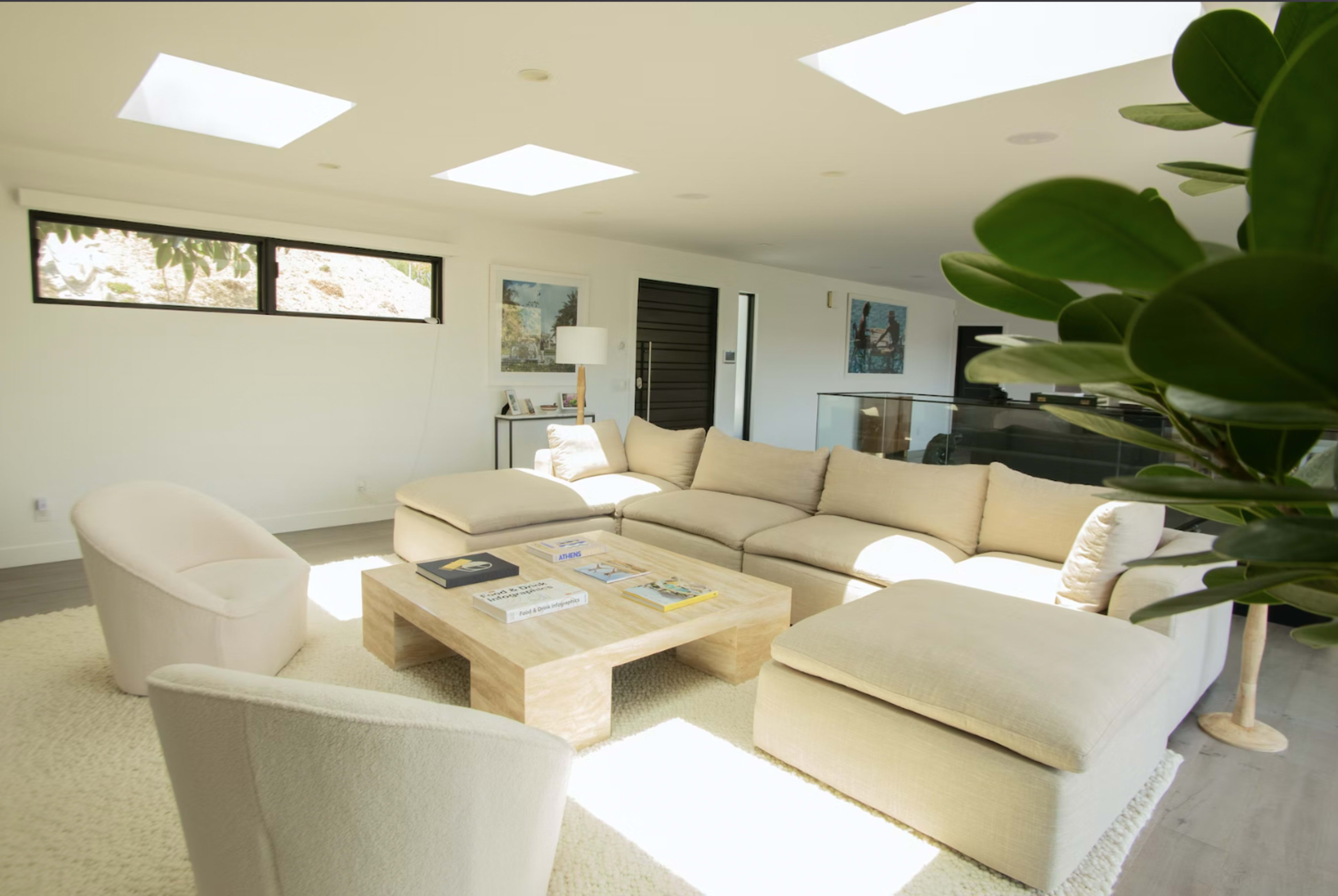 The image shows a modern living room featuring a large beige sectional sofa, a glass coffee table with various magazines, and two accent chairs, illuminated by natural light through skylights.