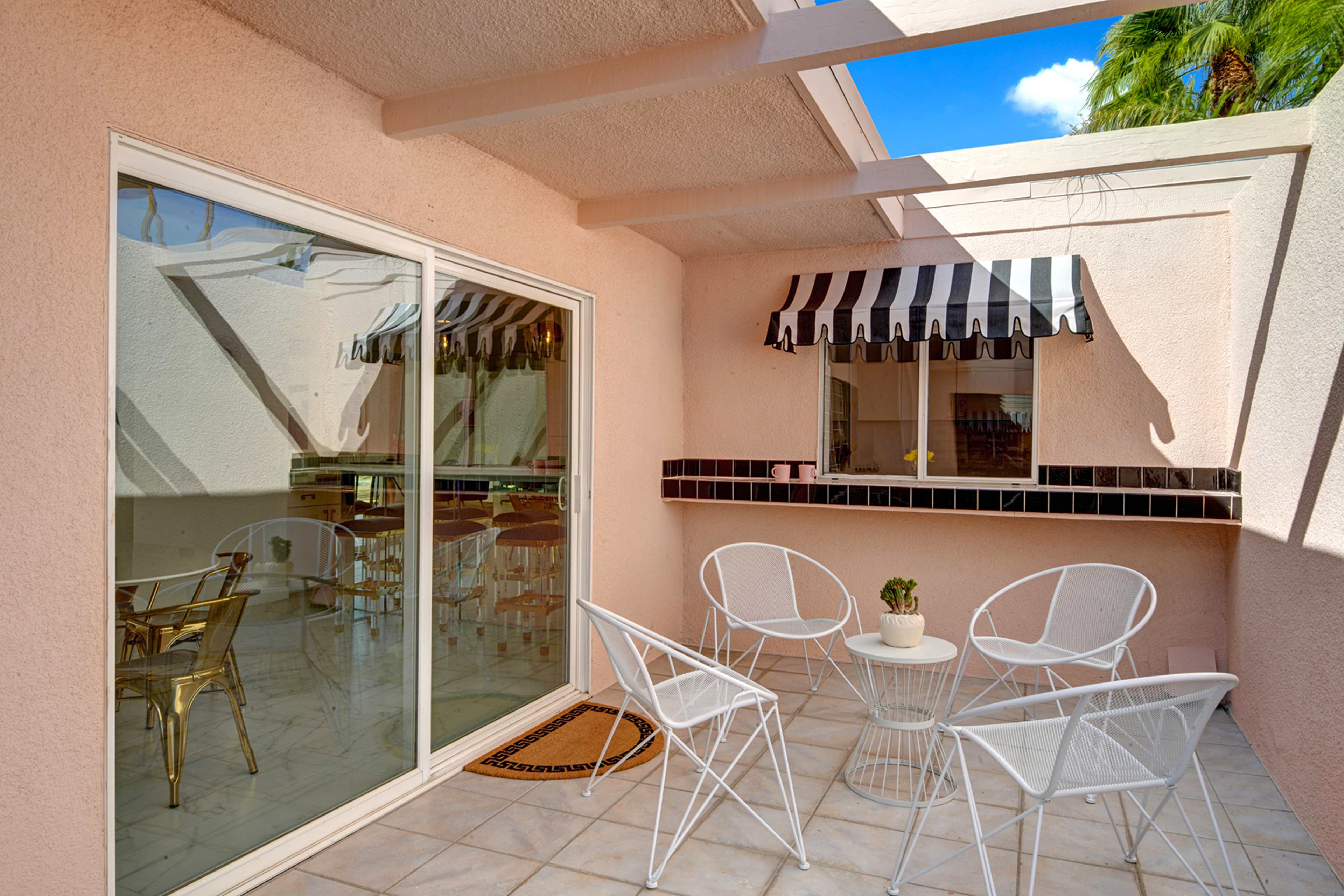 A furnished patio features a glass table surrounded by white metal chairs, with a window and striped awning in the background.