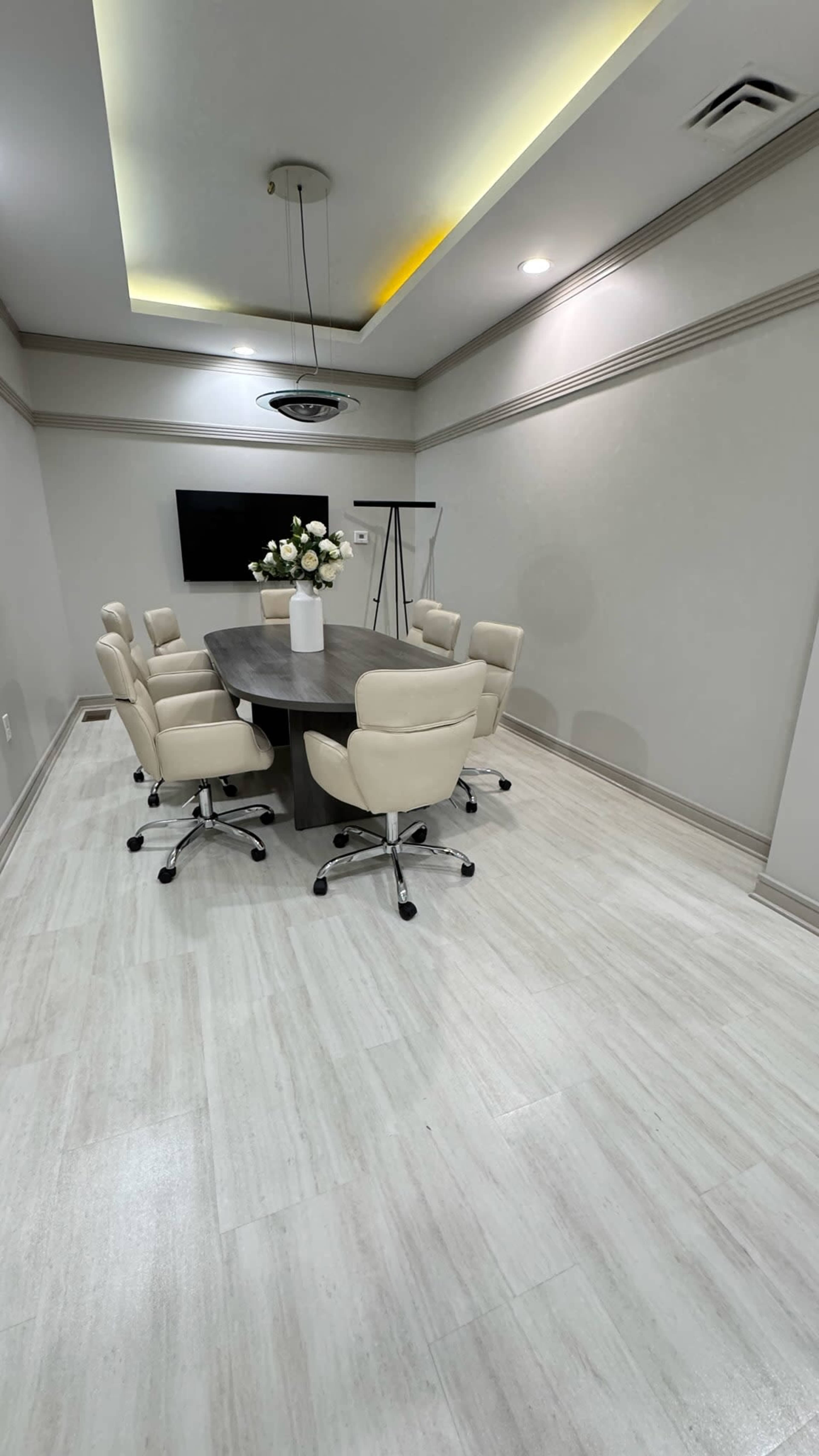 A meeting room features a round table surrounded by six light-colored chairs, with a television, flip chart stand, and a vase of flowers on the table.