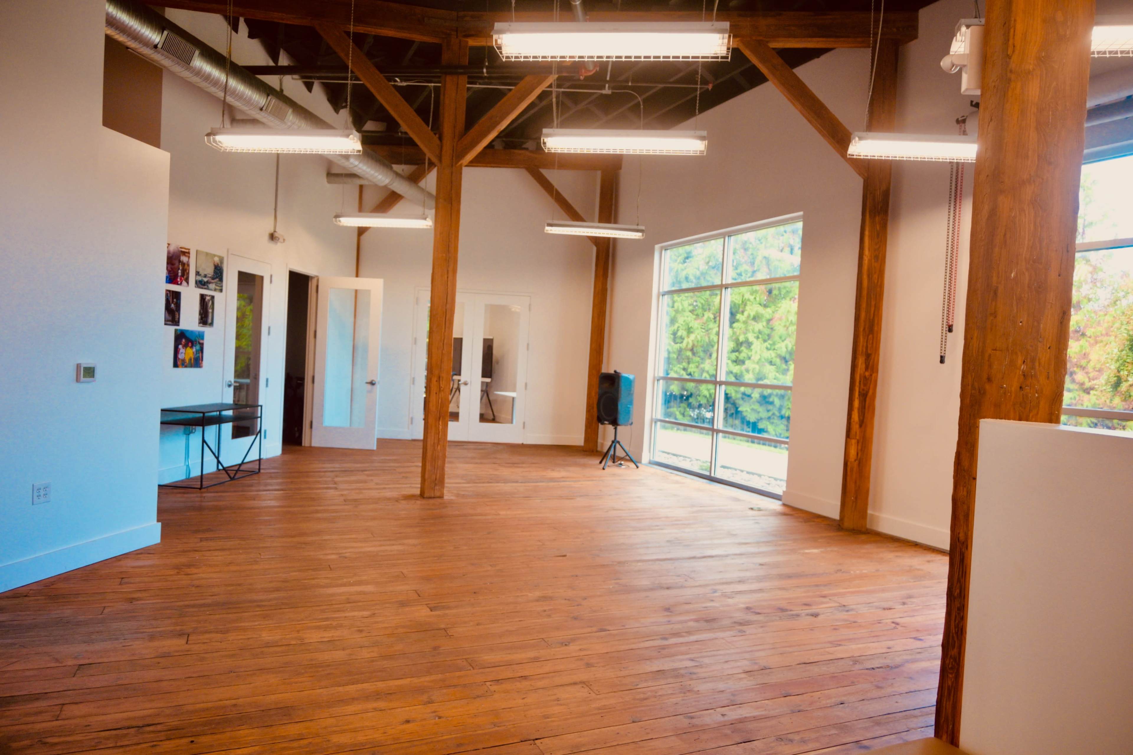 The image shows an empty room with wooden floors, exposed wooden beams, and large windows allowing natural light to fill the space.