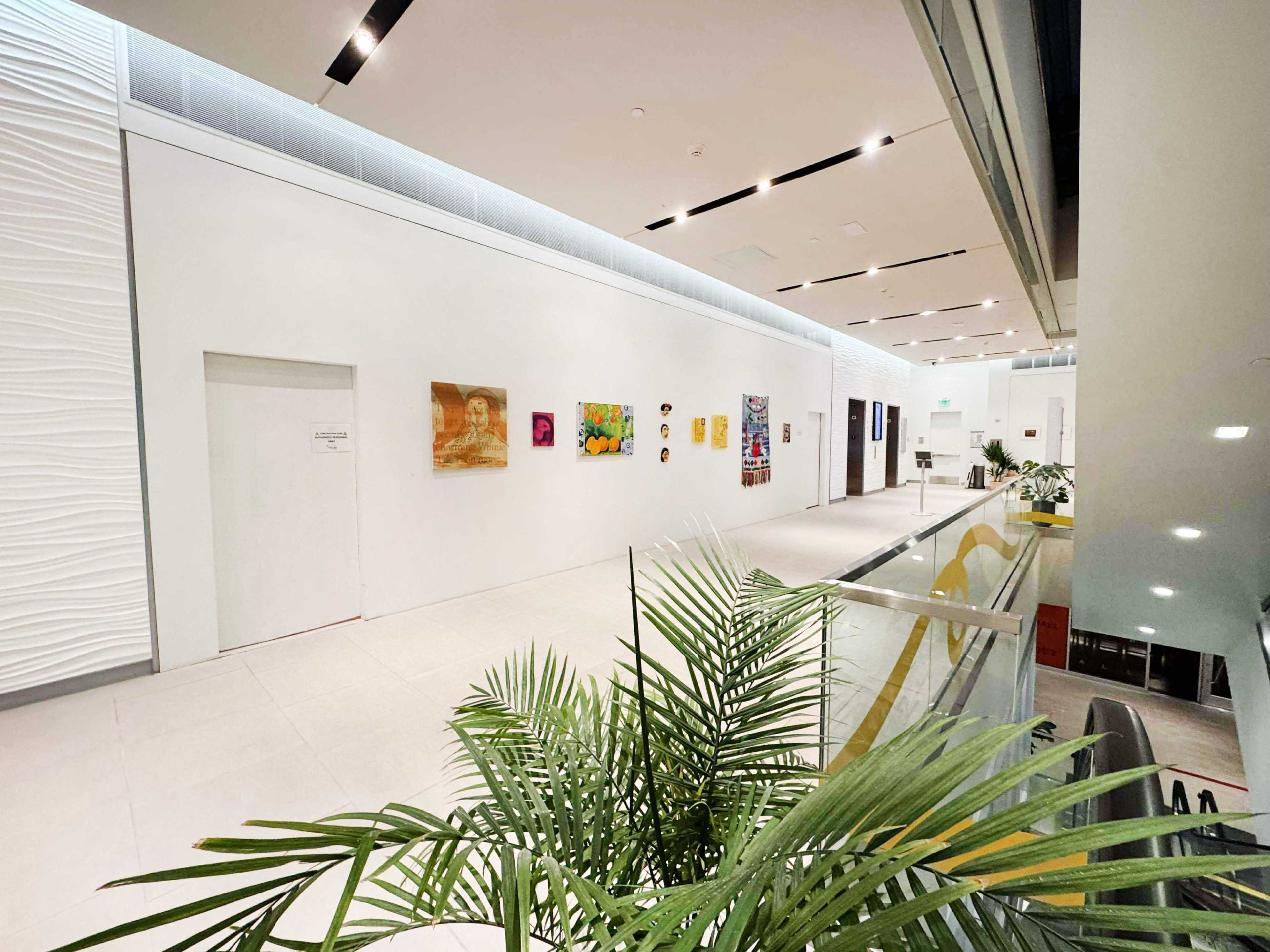 The image shows a modern indoor space with white walls adorned with various pieces of art, a glass railing, and a tropical plant in the foreground.