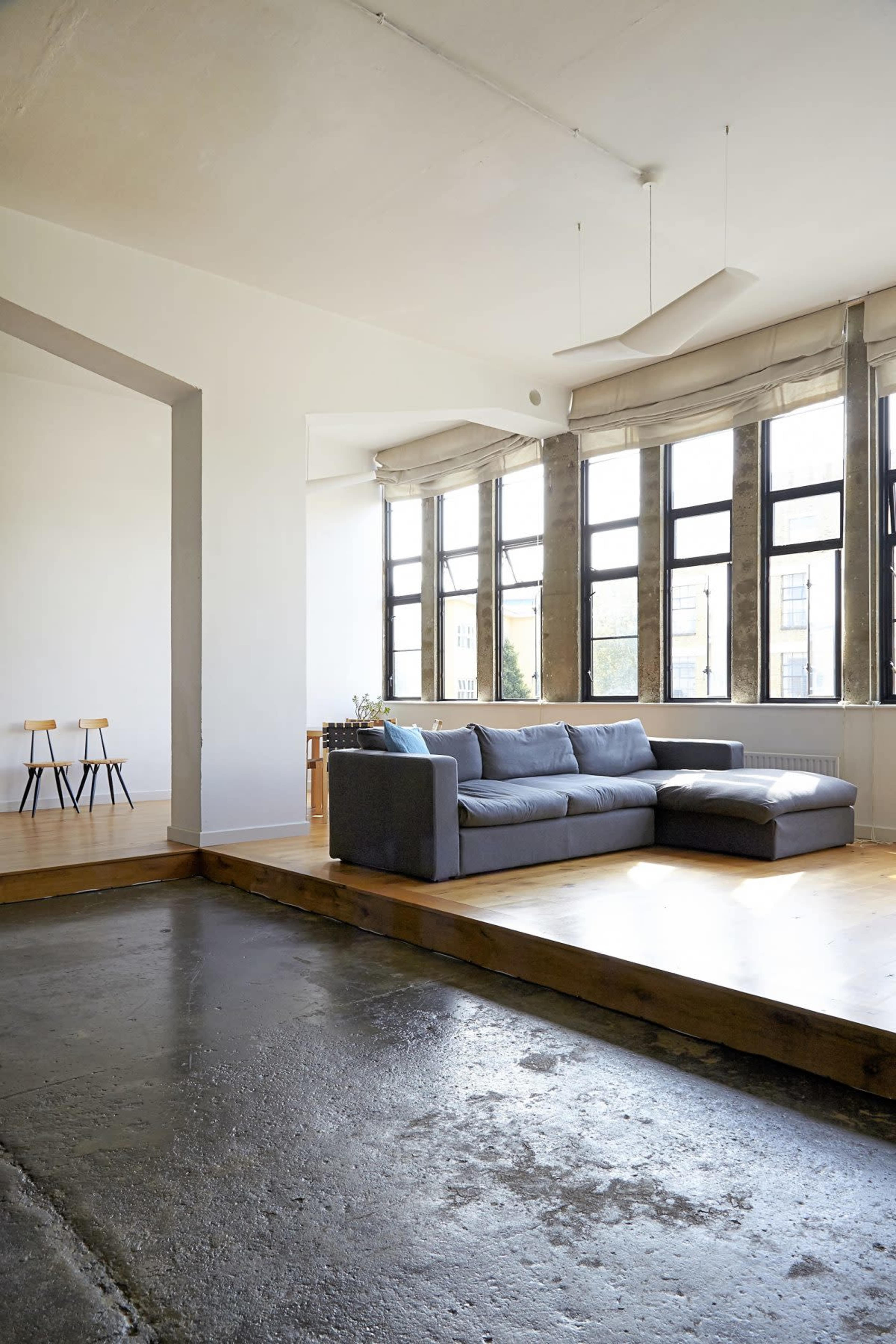 The image shows a spacious living room featuring a large gray sectional couch, wooden flooring, and large windows allowing natural light.