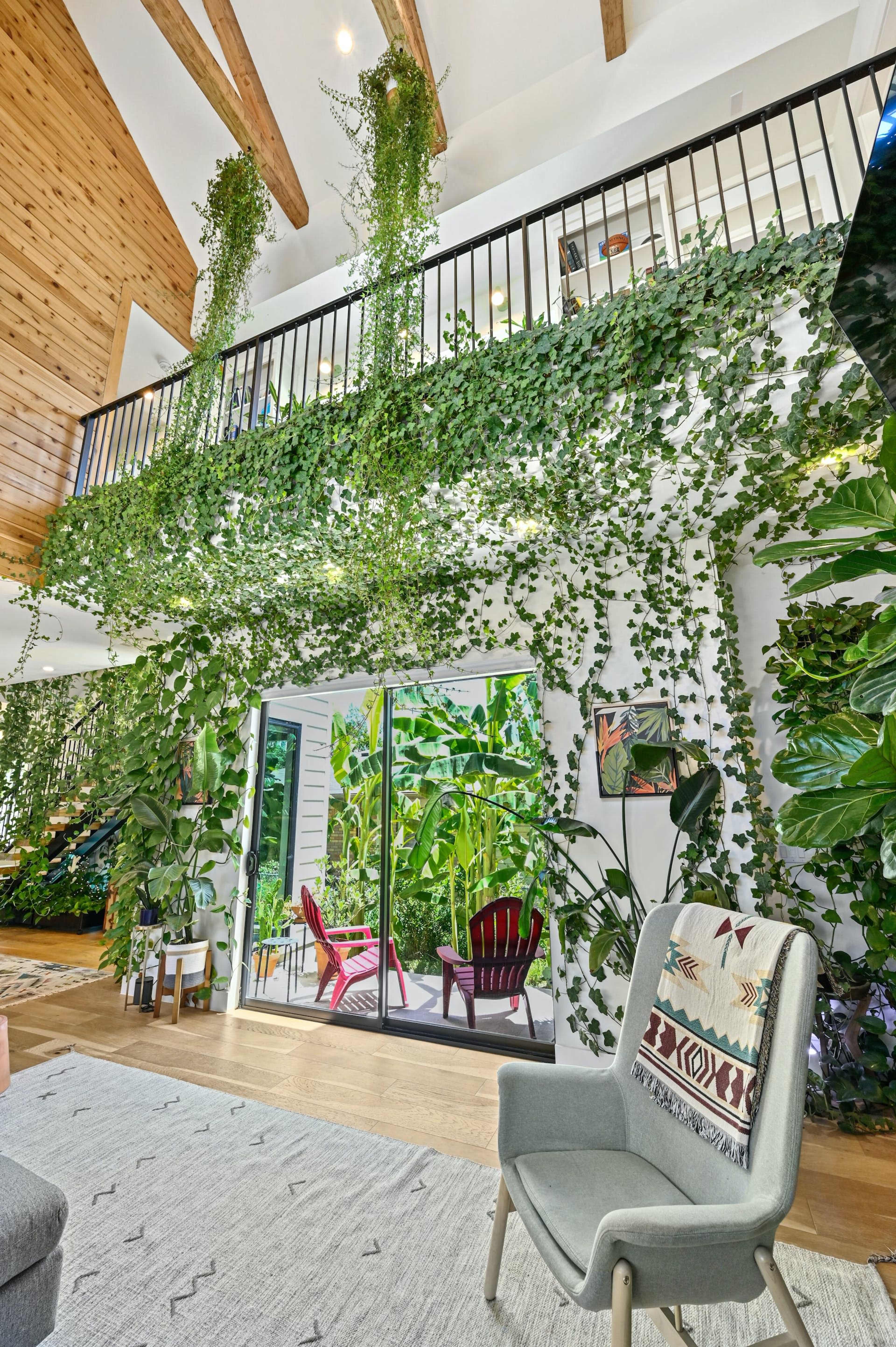 The interior of a brightly lit, two-story living room features tall plants cascading from the balcony and large sliding glass doors leading to a garden patio.