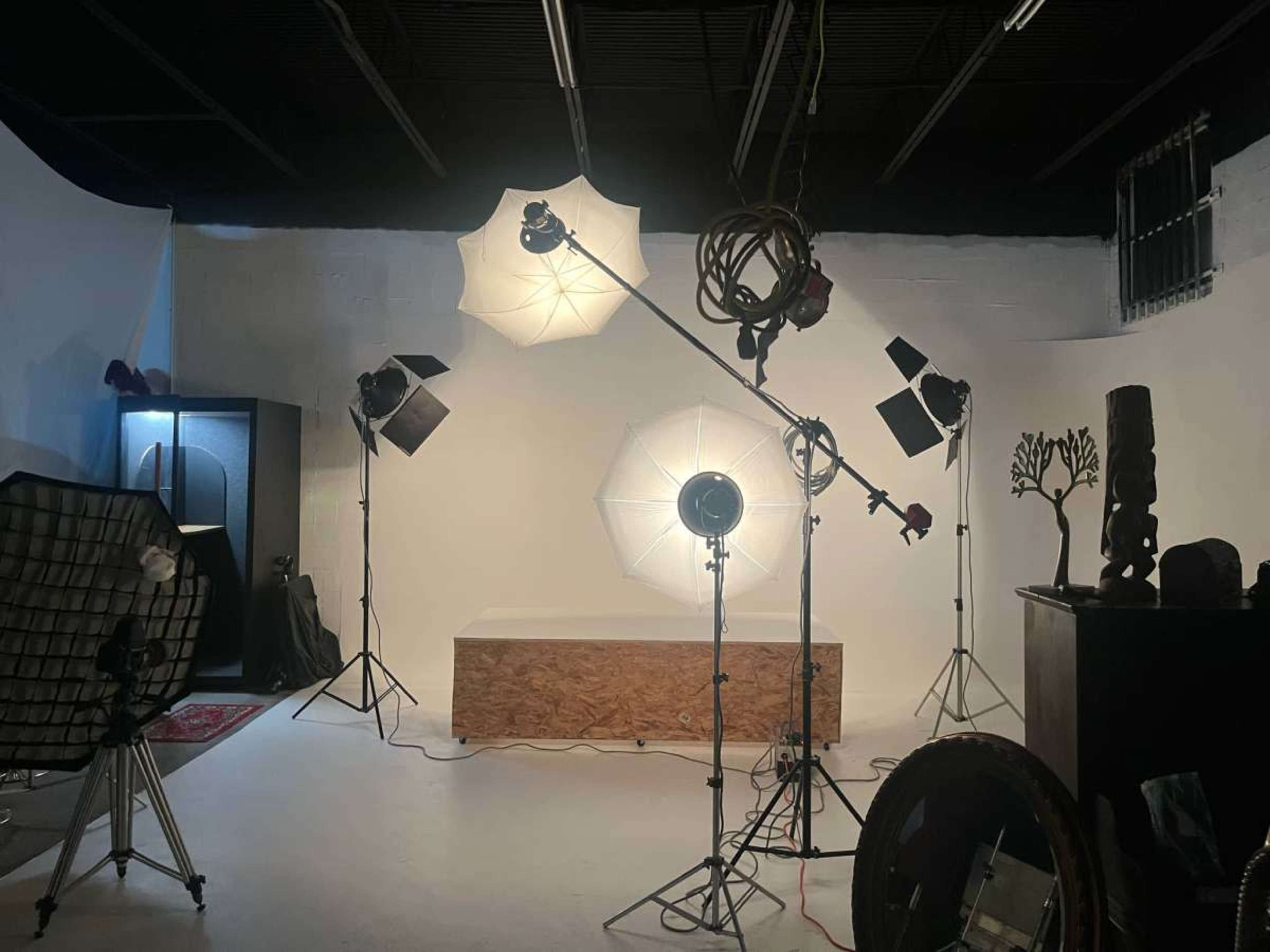 The image shows a photography studio with various lighting equipment set up around a wooden platform against a plain white backdrop.