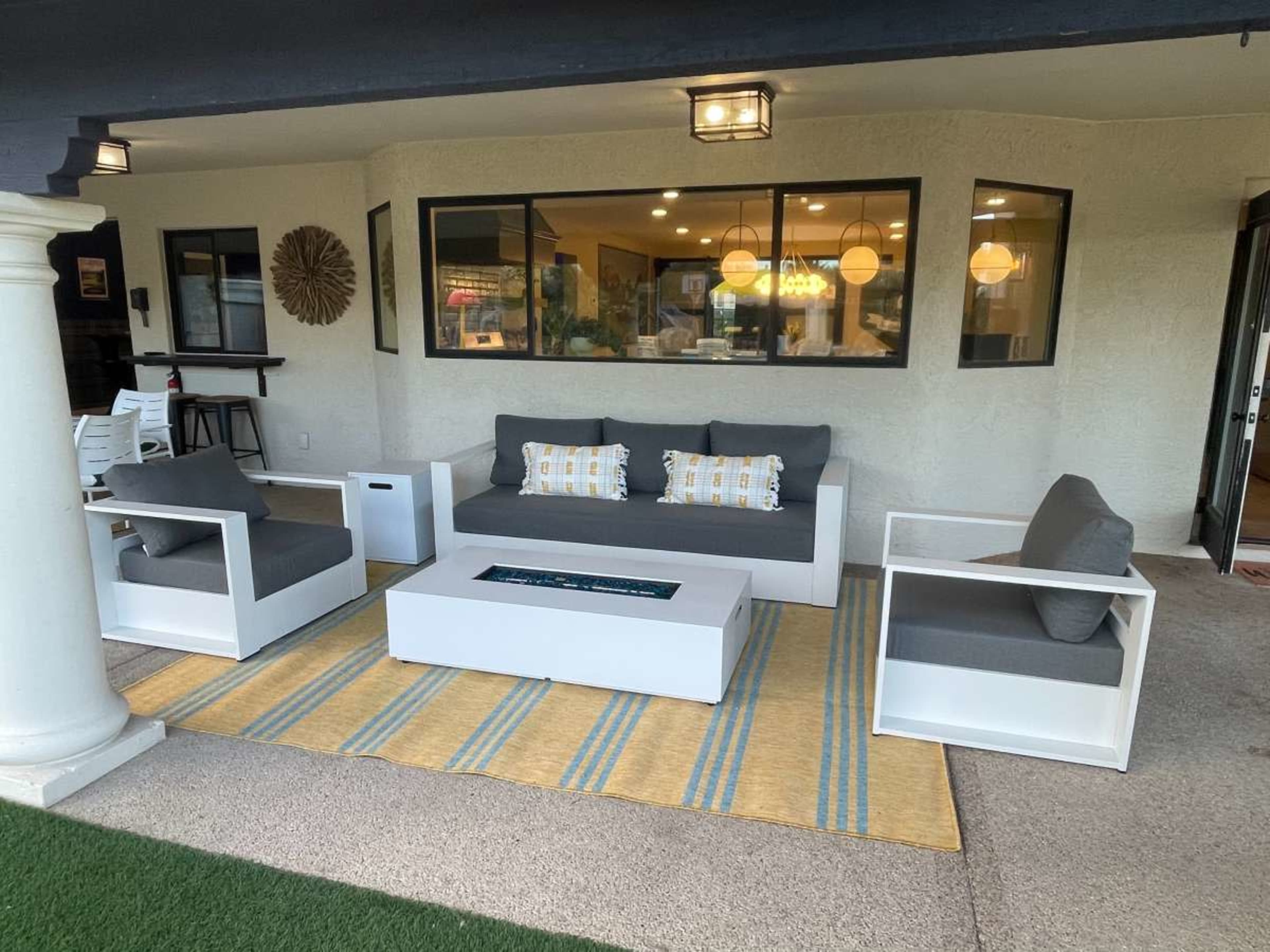The image shows a modern outdoor seating area with a gray sofa, armchairs, and a rectangular coffee table, arranged on a striped rug in front of a house.