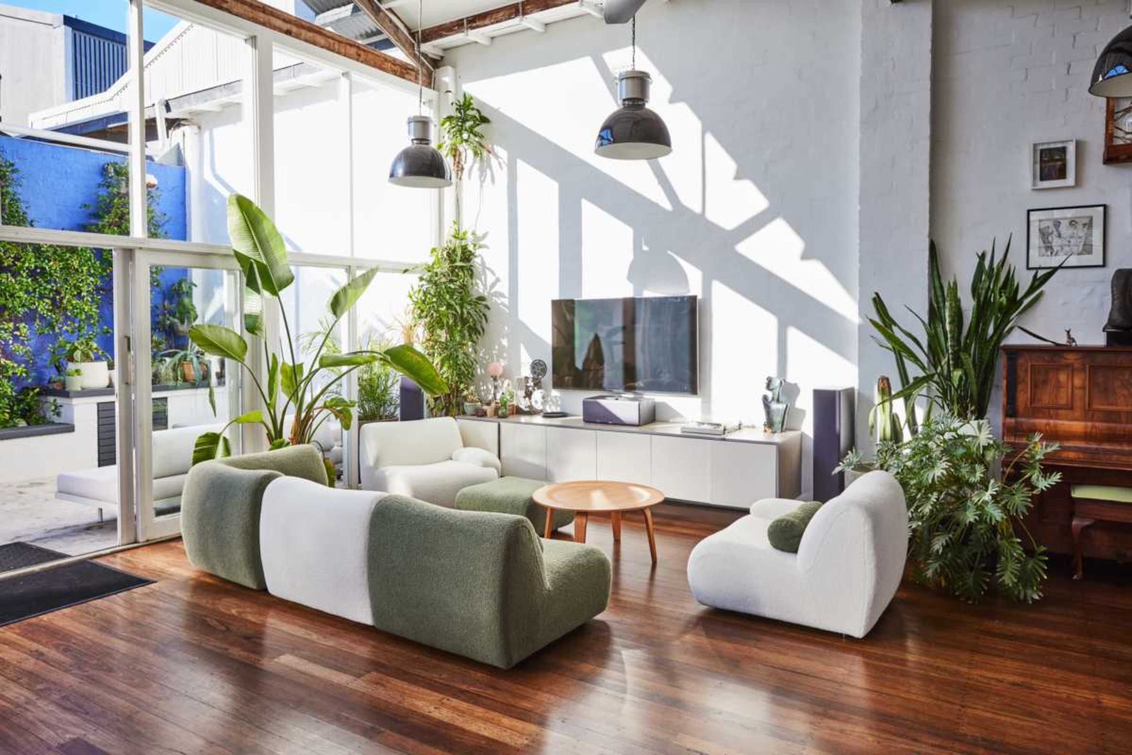 The image shows a modern living room with large windows, featuring green and white furniture, a flat-screen TV, and various plants.