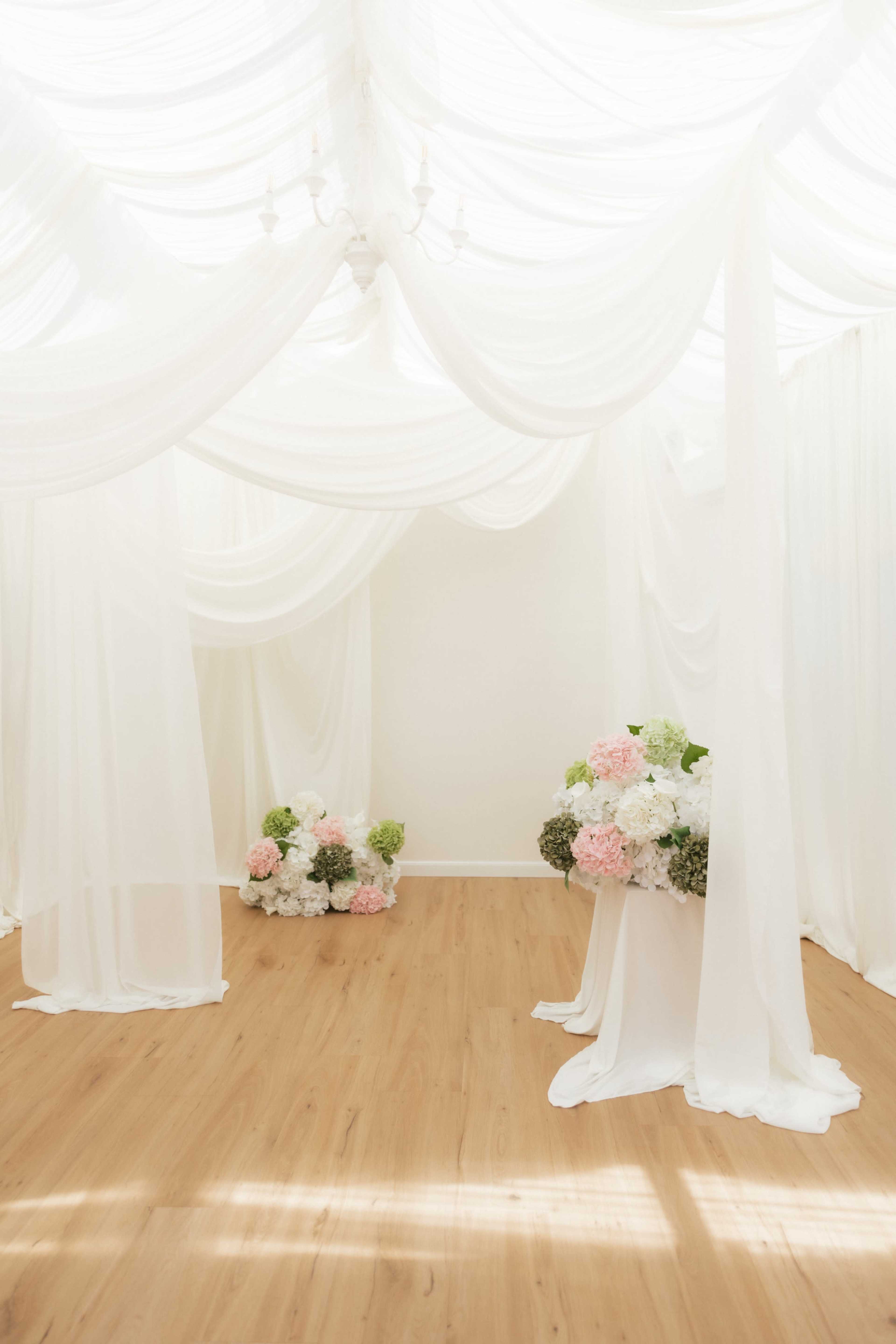 A bright event space features draped white fabric along the ceiling and walls, with floral arrangements in soft colors positioned on the floor.