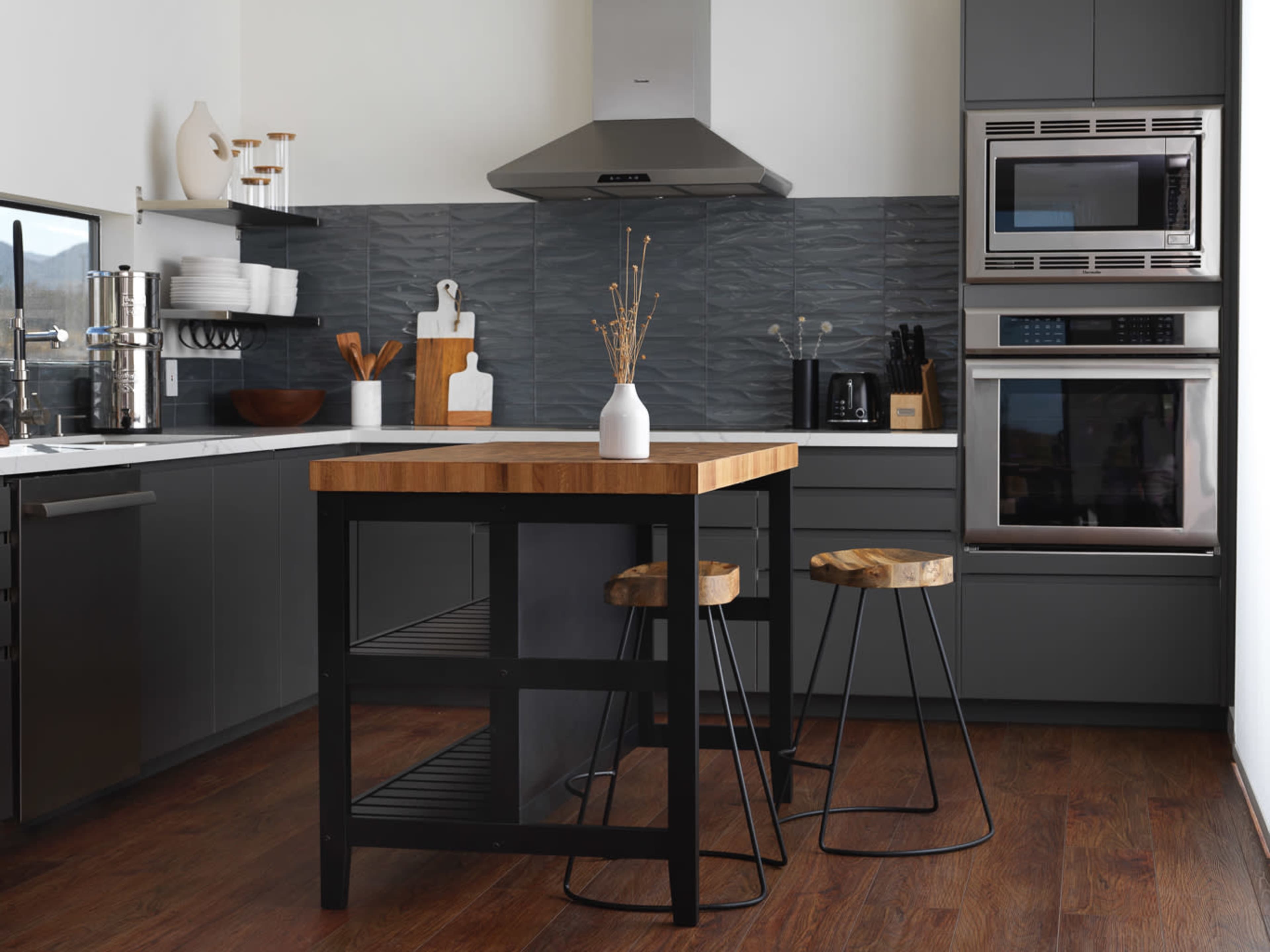 The modern kitchen features a central wooden island with two metal stools, gray cabinetry, stainless steel appliances, and a textured backsplash.