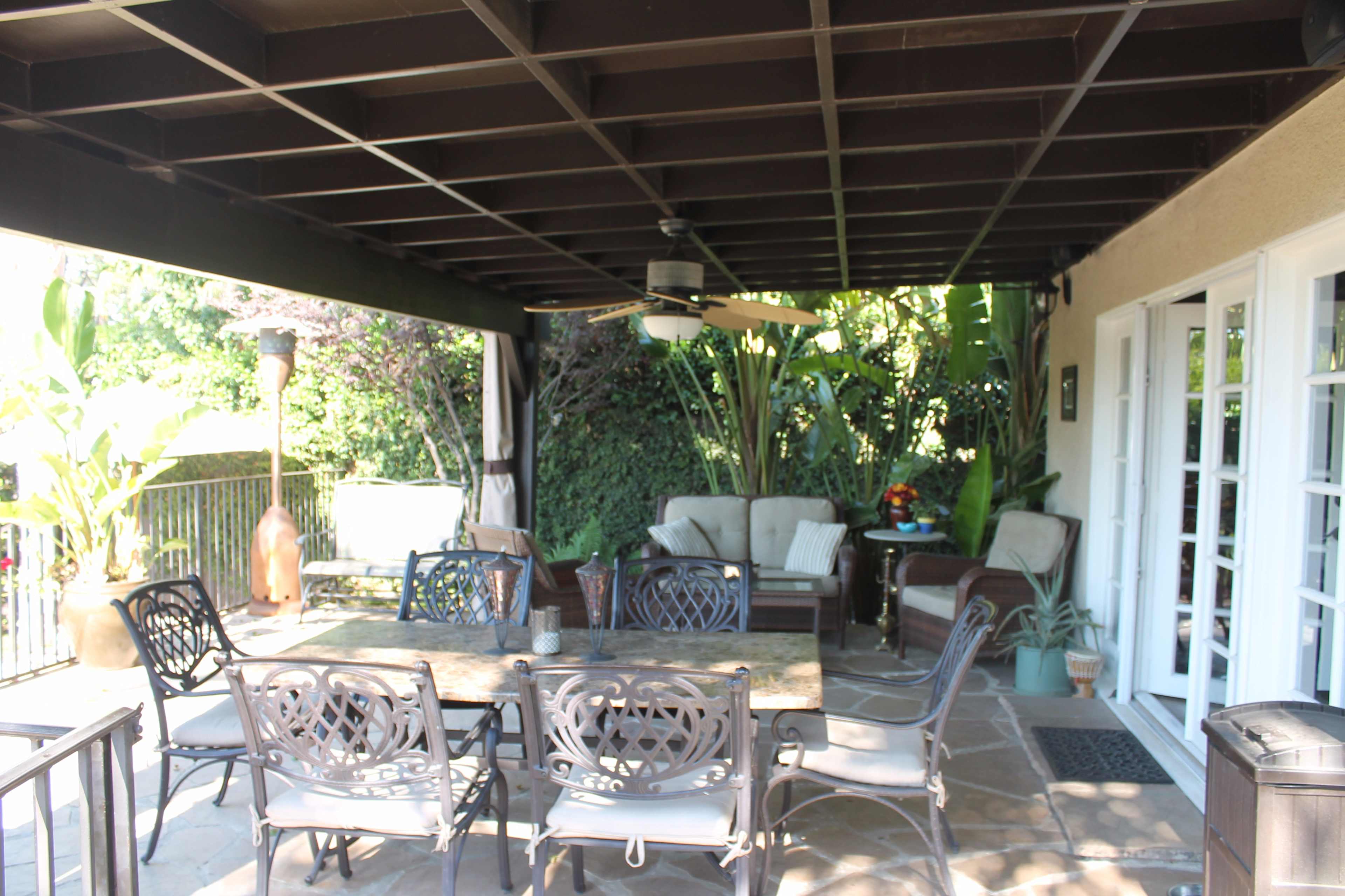 The image shows a covered patio area furnished with metal chairs and a table, surrounded by greenery.